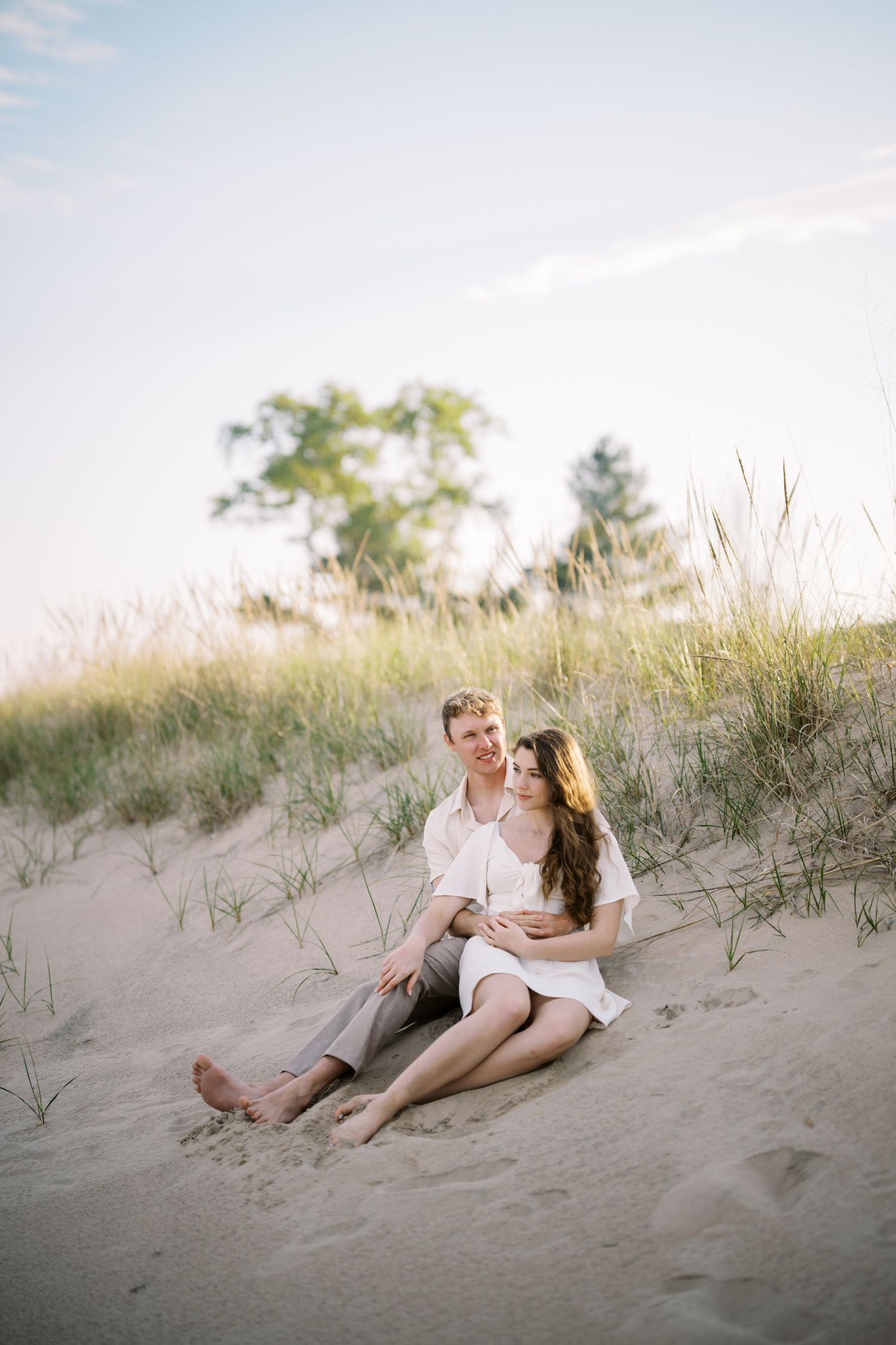 A romantic couple sits together on sandy dunes during a dreamy sunset photoshoot at the beach.