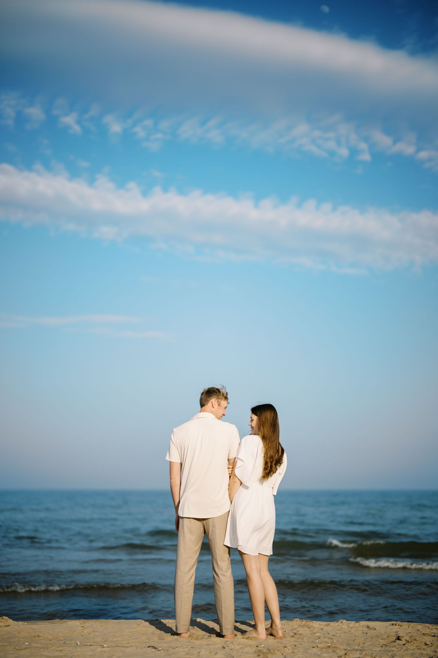 A couple in light summer attire stands together on a sandy beach looking out at the ocean under wispy clouds.
