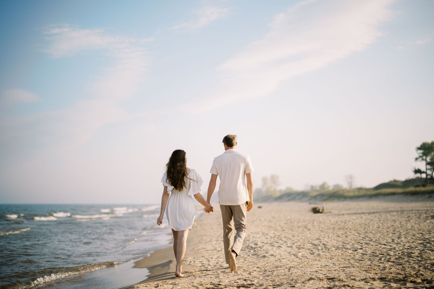 Couple walking hand in hand on a sandy beach at sunset with ocean waves and blue sky in the background.