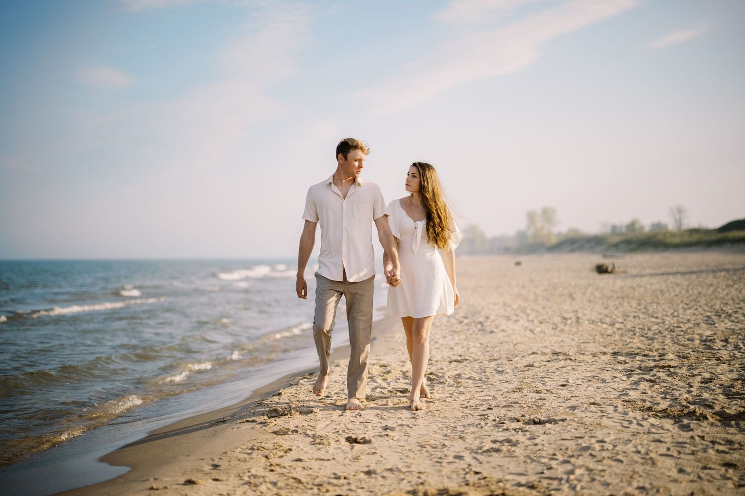 A couple in white clothing walking together along a sandy beach at sunset with waves washing up on shore.