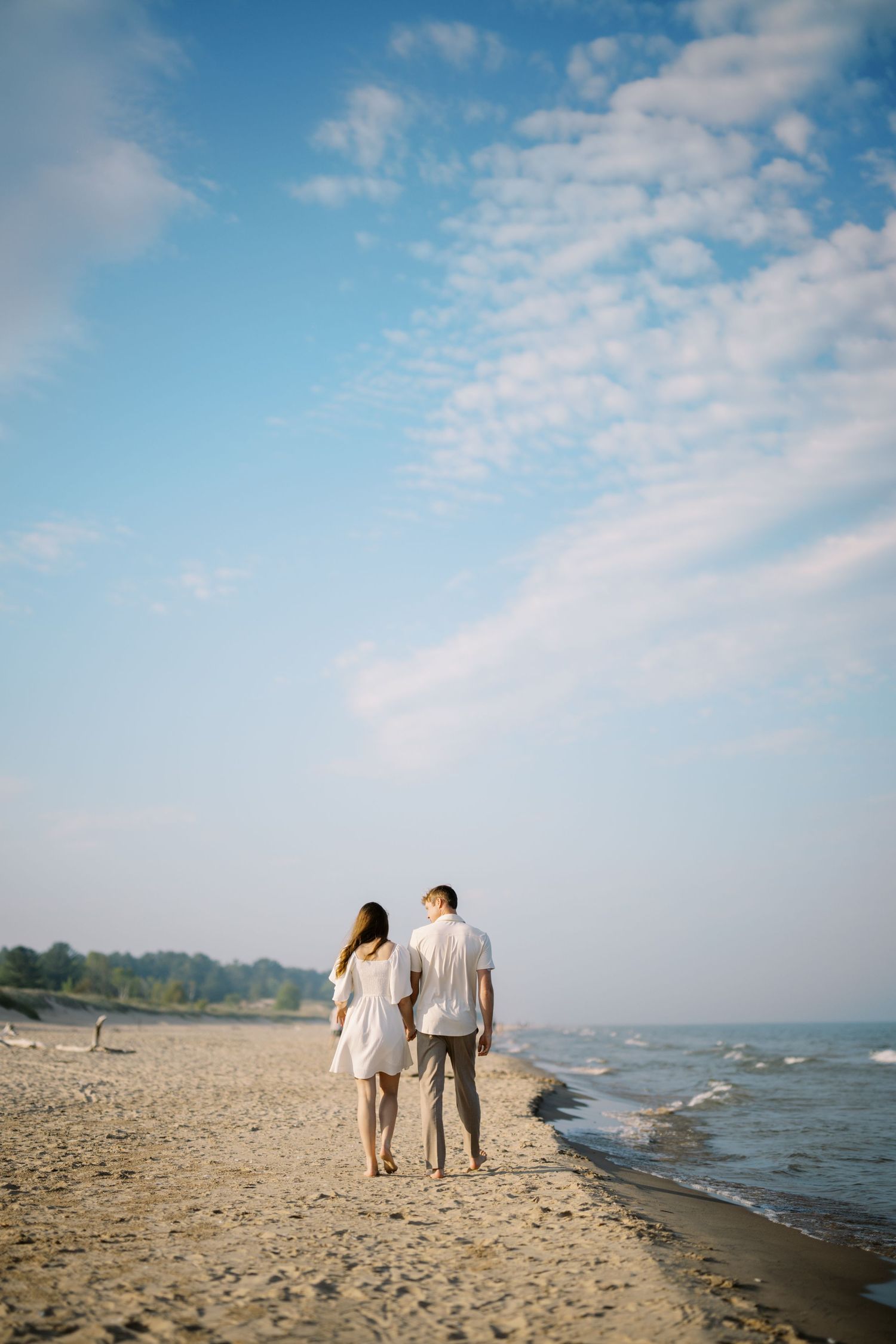 A couple stands together on a serene beach shoreline under wispy clouds and blue sky.