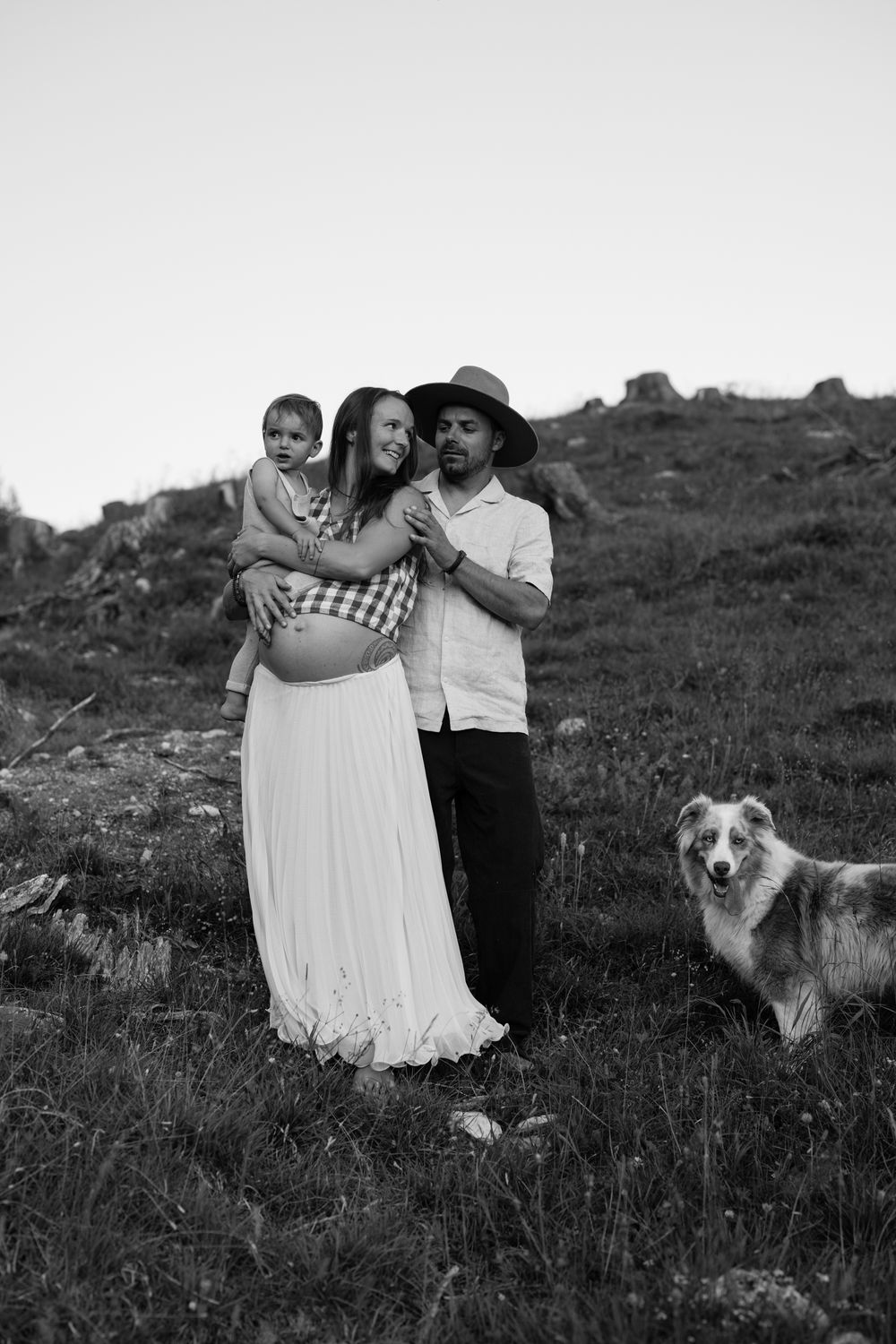 A black and white series of photos showing a couple embracing on a grassy hillside with their dog watching nearby.