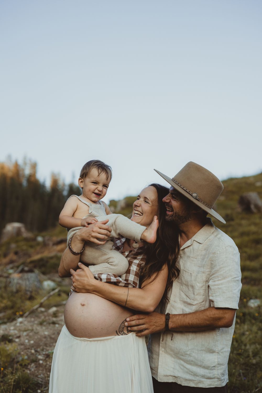 An expectant couple shares a joyful moment outdoors with their baby against a scenic mountain backdrop at sunset.