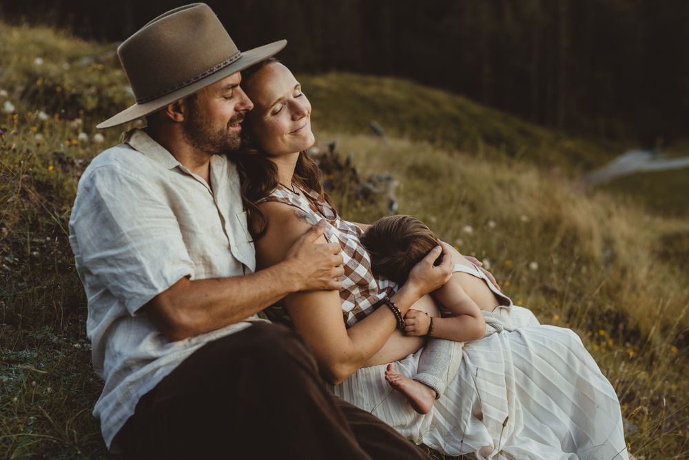 Couple sitting together in rustic outdoor setting at sunset holding glass of wine while relaxing on grassy hillside.