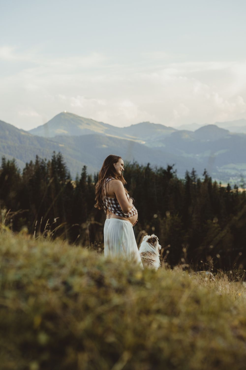 A woman in a flowing white skirt stands with her small dog on a mountaintop overlooking a scenic valley at sunset.