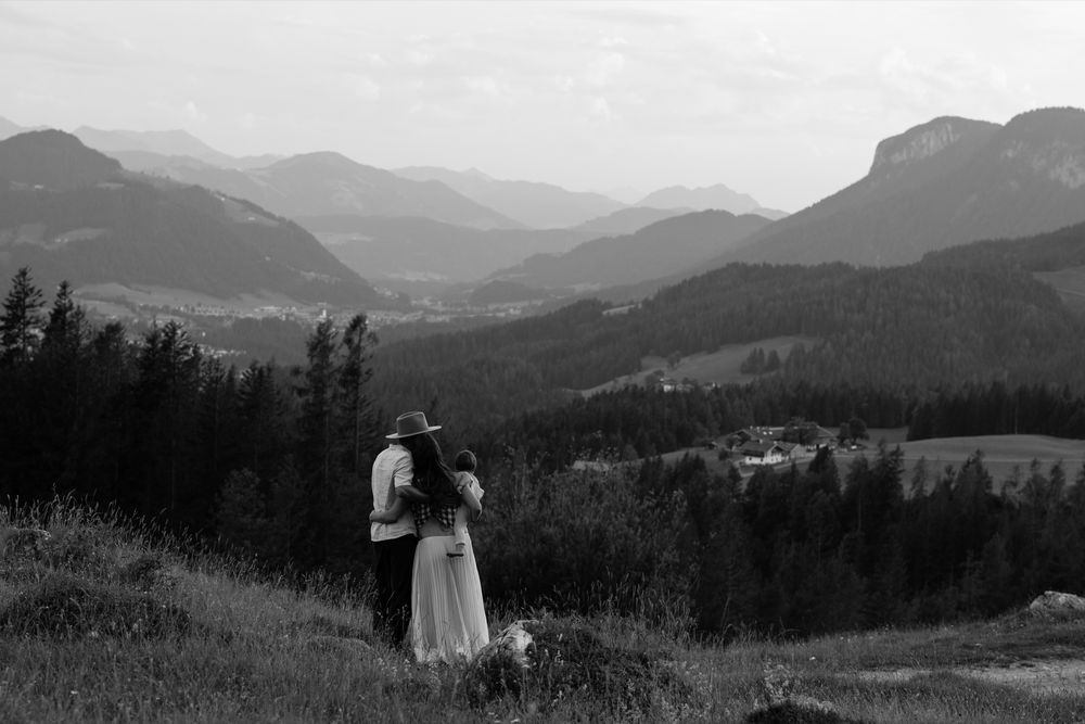 A couple embraces while overlooking a scenic mountain landscape with rolling hills and forest in black and white.