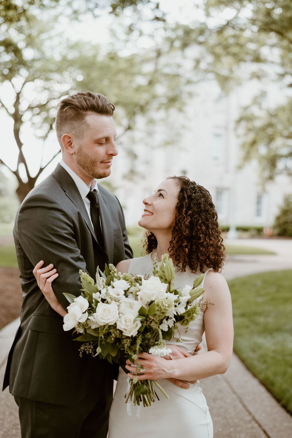 A romantic wedding couple embraces outdoors while holding a white and green floral bouquet against a soft natural background.
