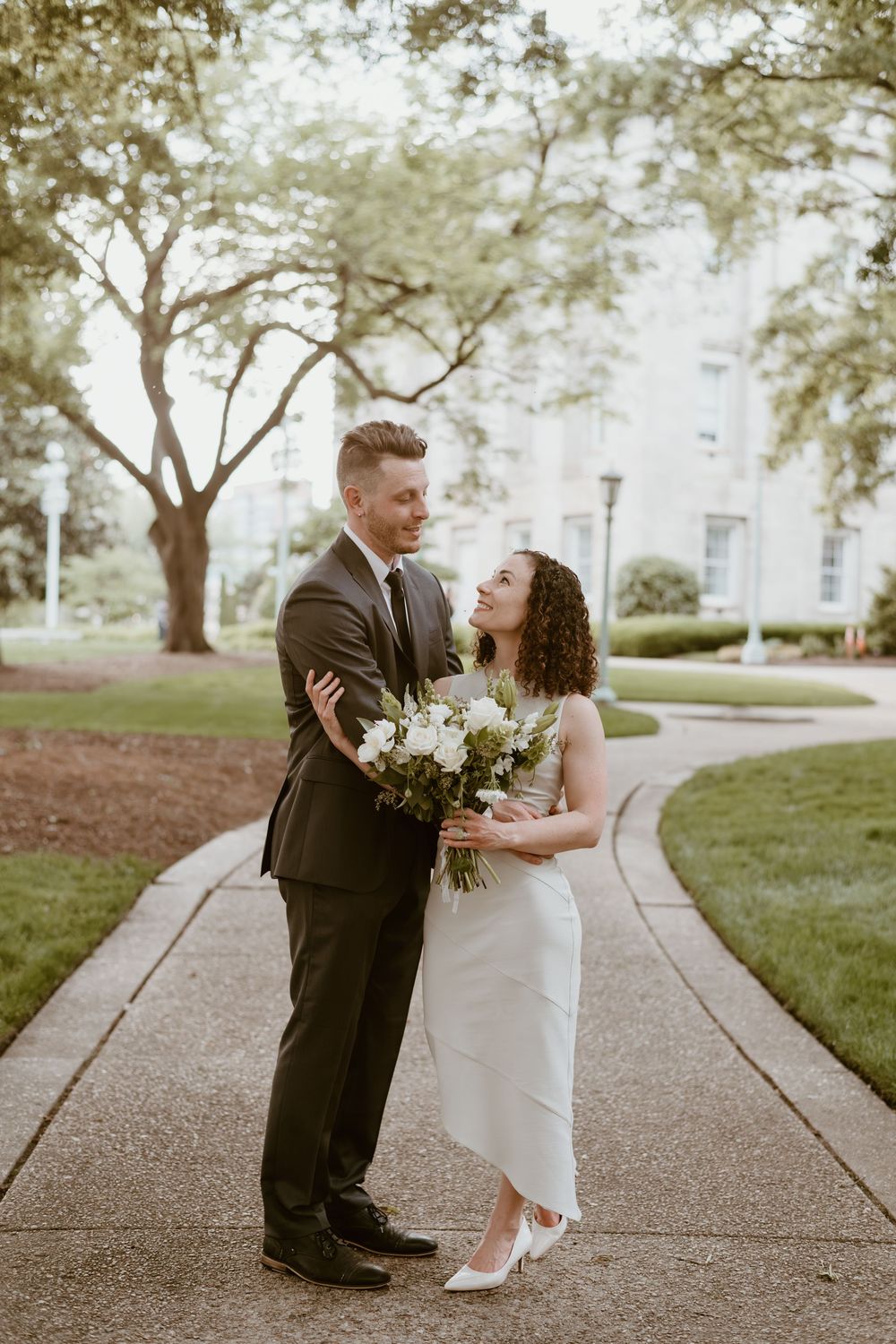 A couple stands together on a winding garden path holding a white floral bouquet while celebrating their special day.