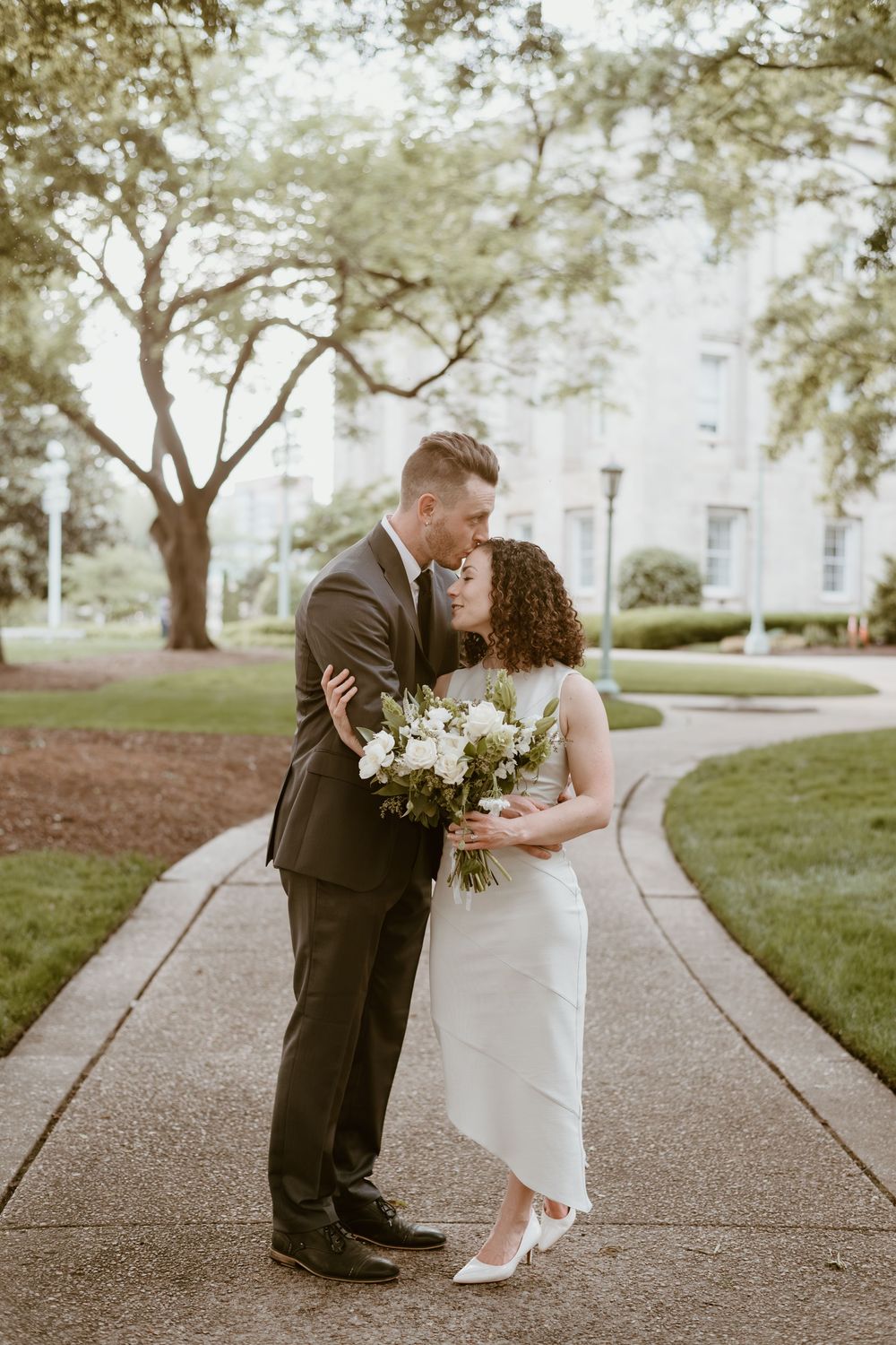 A couple shares an intimate moment on a tree-lined pathway while holding a white floral bouquet.