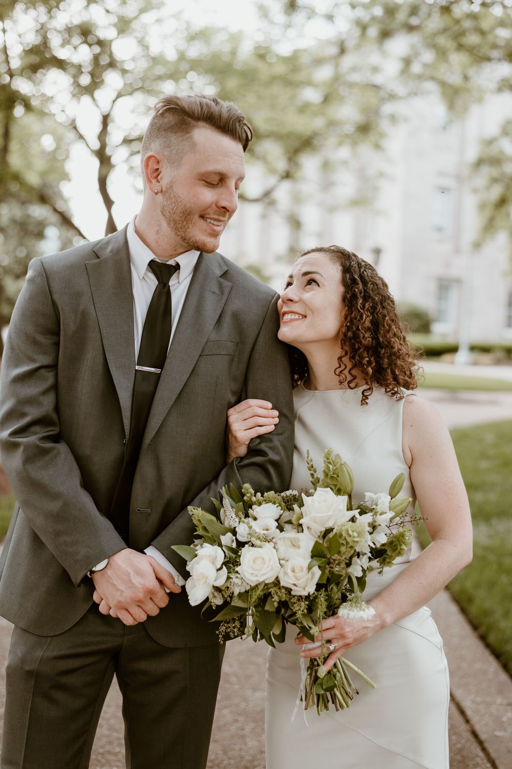Joyful newlyweds share a tender moment outside as the bride holds a beautiful white floral bouquet in her sleeveless dress.