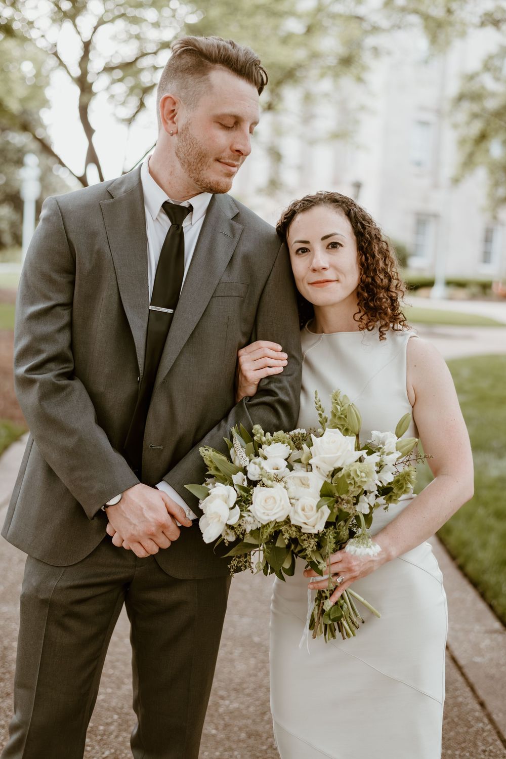 Elegantly dressed couple poses outdoors with white floral bouquet at their intimate garden wedding ceremony.