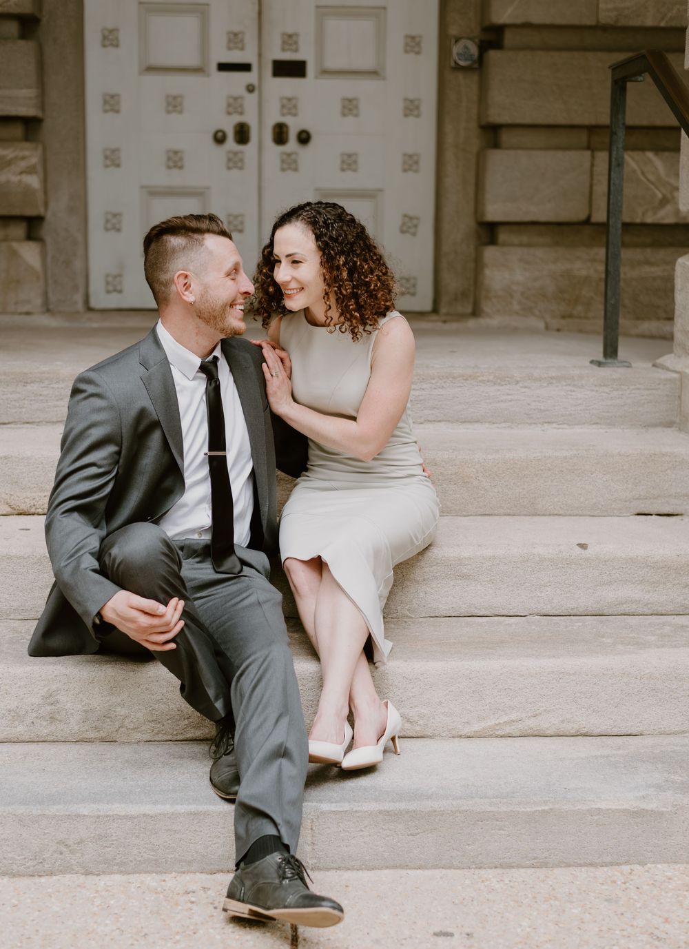 Stylishly dressed couple sitting together on stone steps sharing an intimate moment in front of vintage doors.