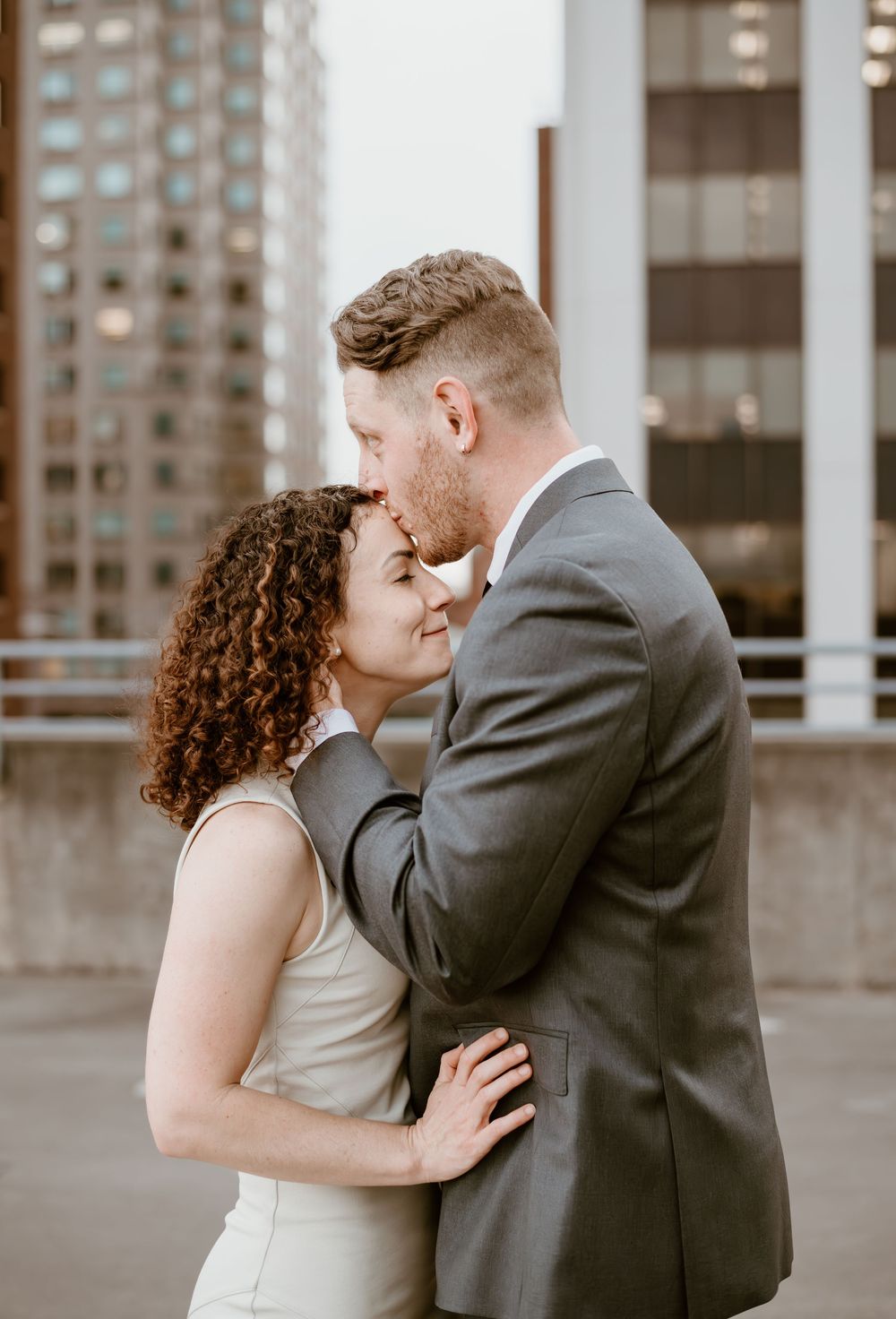 A couple shares an intimate moment on a rooftop, with the bride in white and groom in grey suit against city buildings.