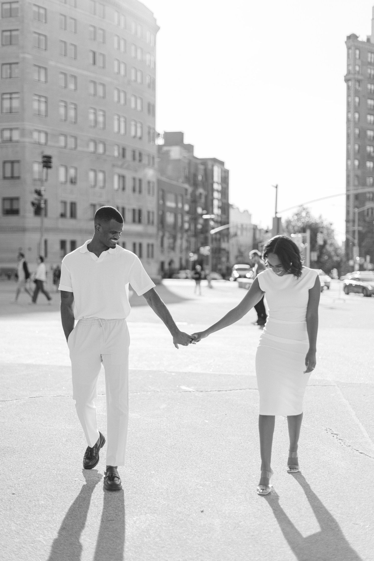 A couple in white outfits holds hands while walking down a city street.