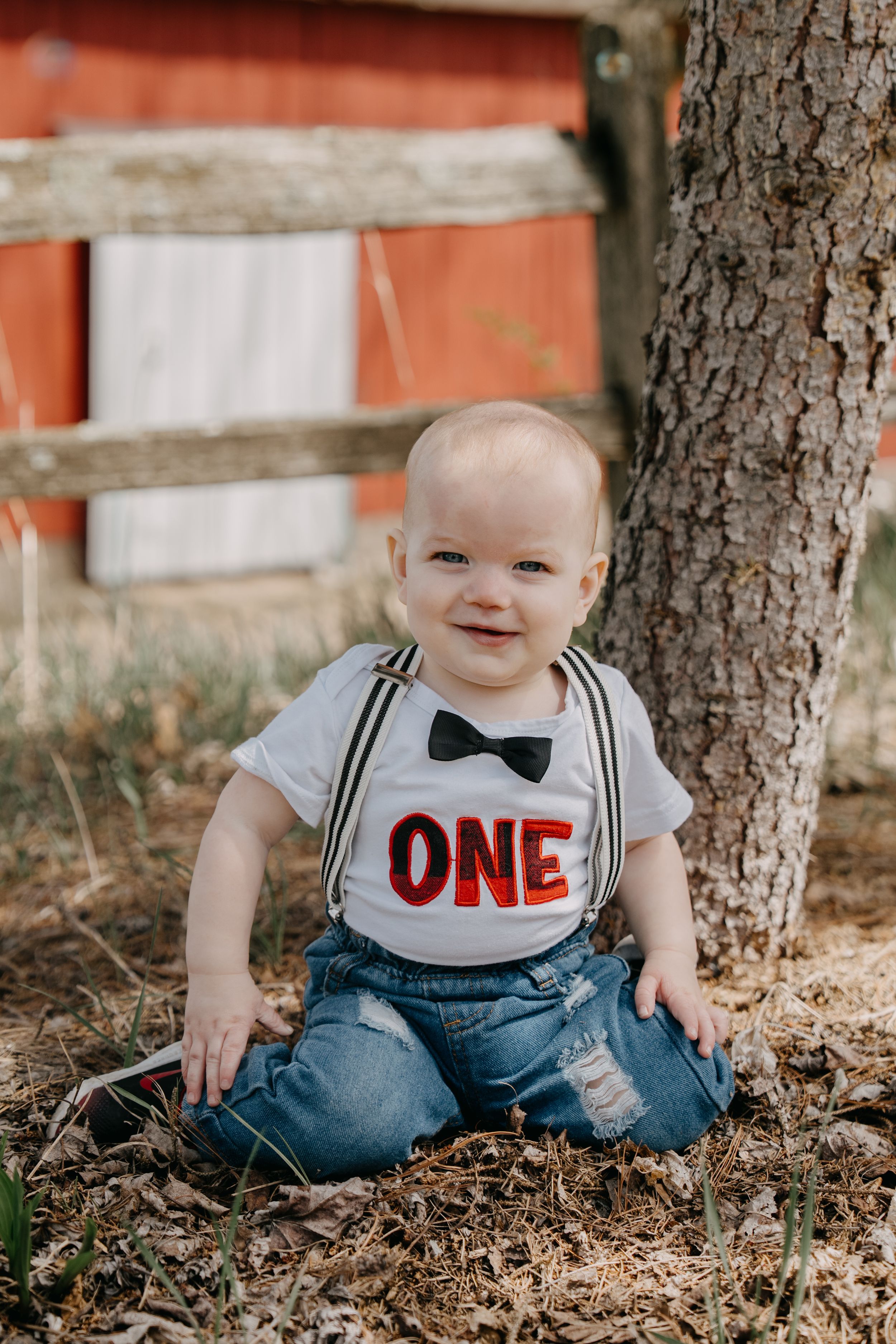 A baby wearing a white bib with "ONE" and a bowtie sits by a tree in distressed jeans.