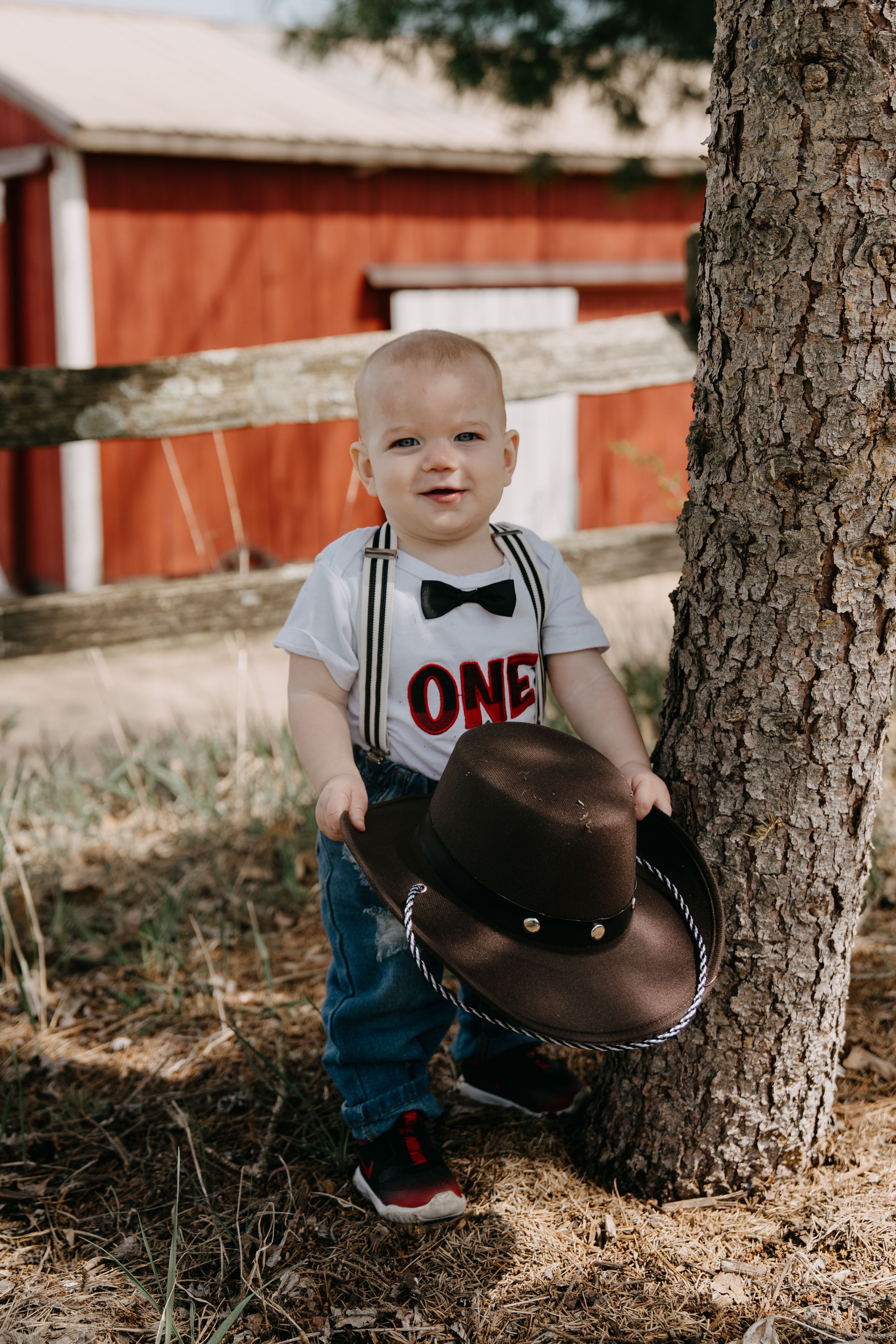 A young toddler in suspenders and bowtie stands next to a tree by a red barn holding a brown cowboy hat.