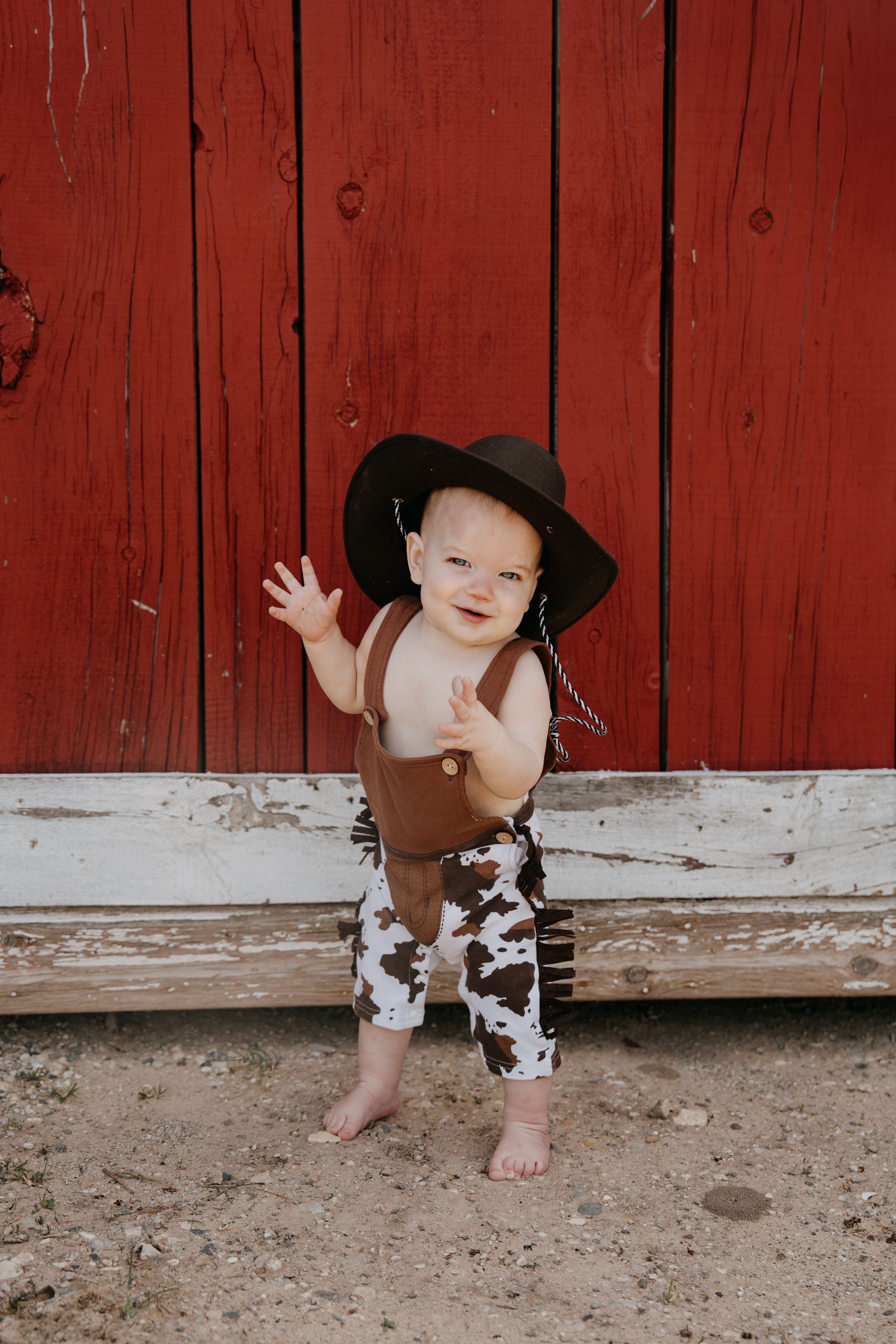 Small child in brown overalls and black hat stands barefoot against rustic red barn wall while waving.