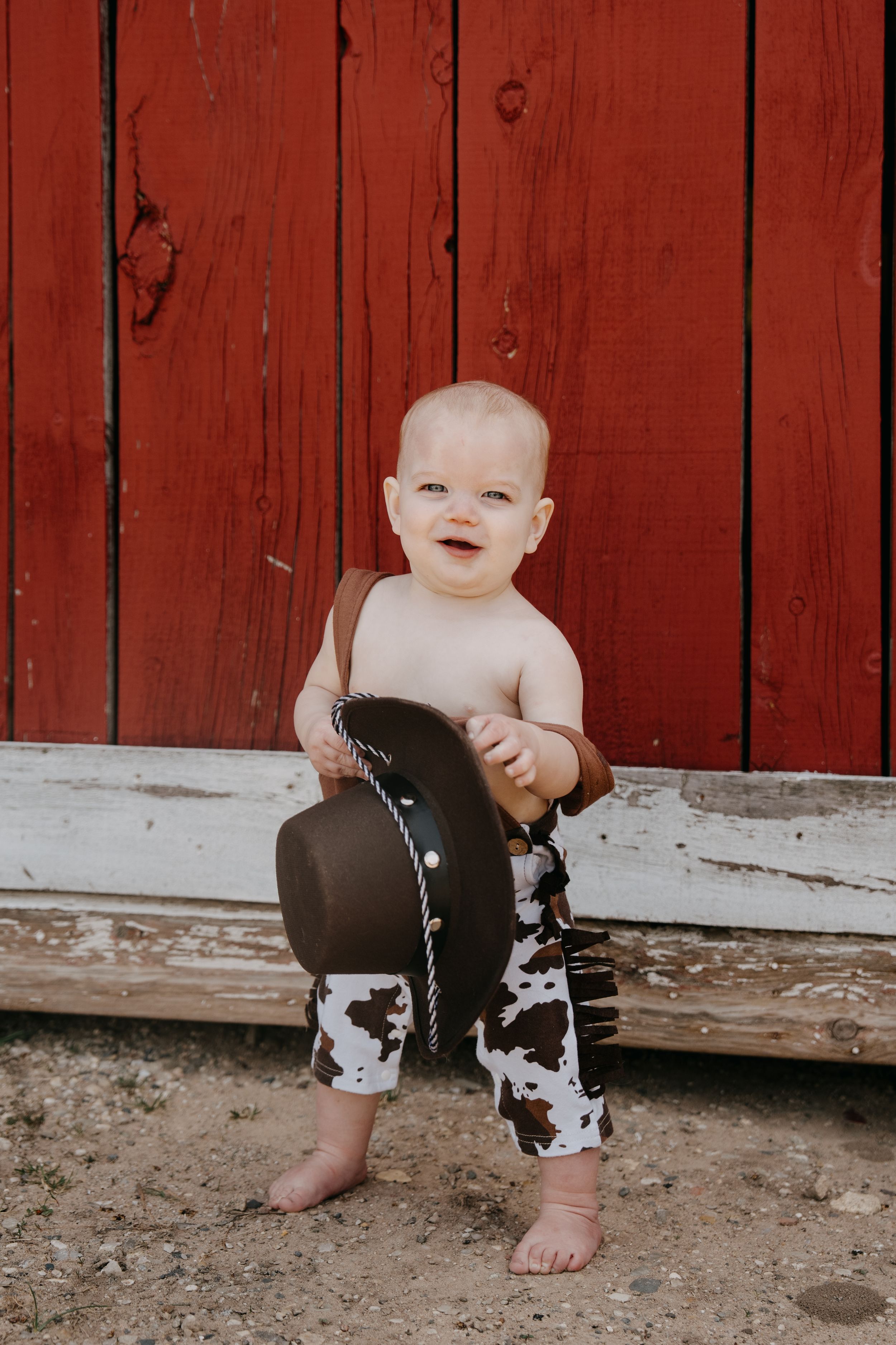 A smiling baby in cow print pants poses against a rustic red barn wall.