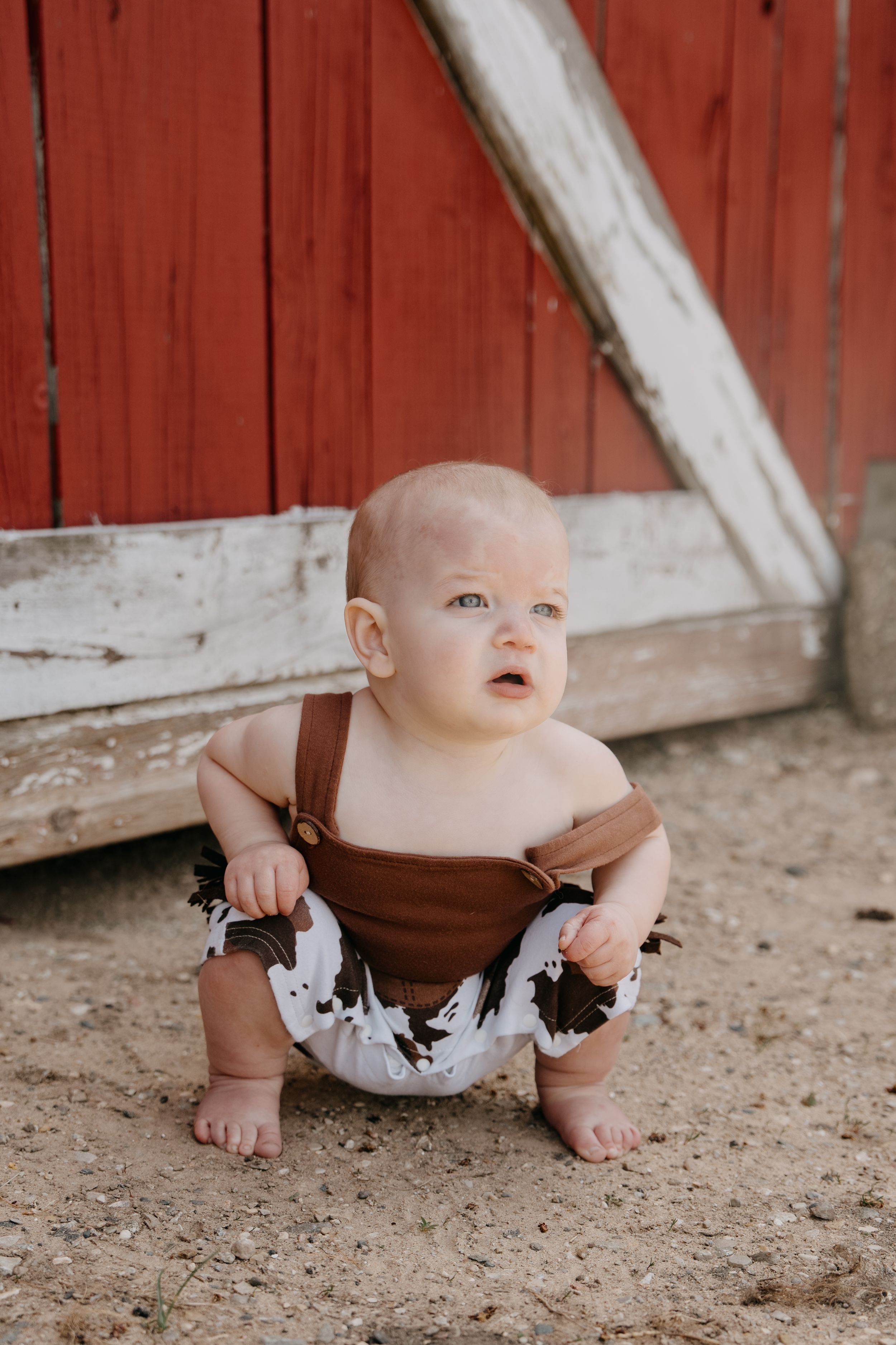 Small child wearing brown and white cow print romper sits on dirt ground against red barn wall.