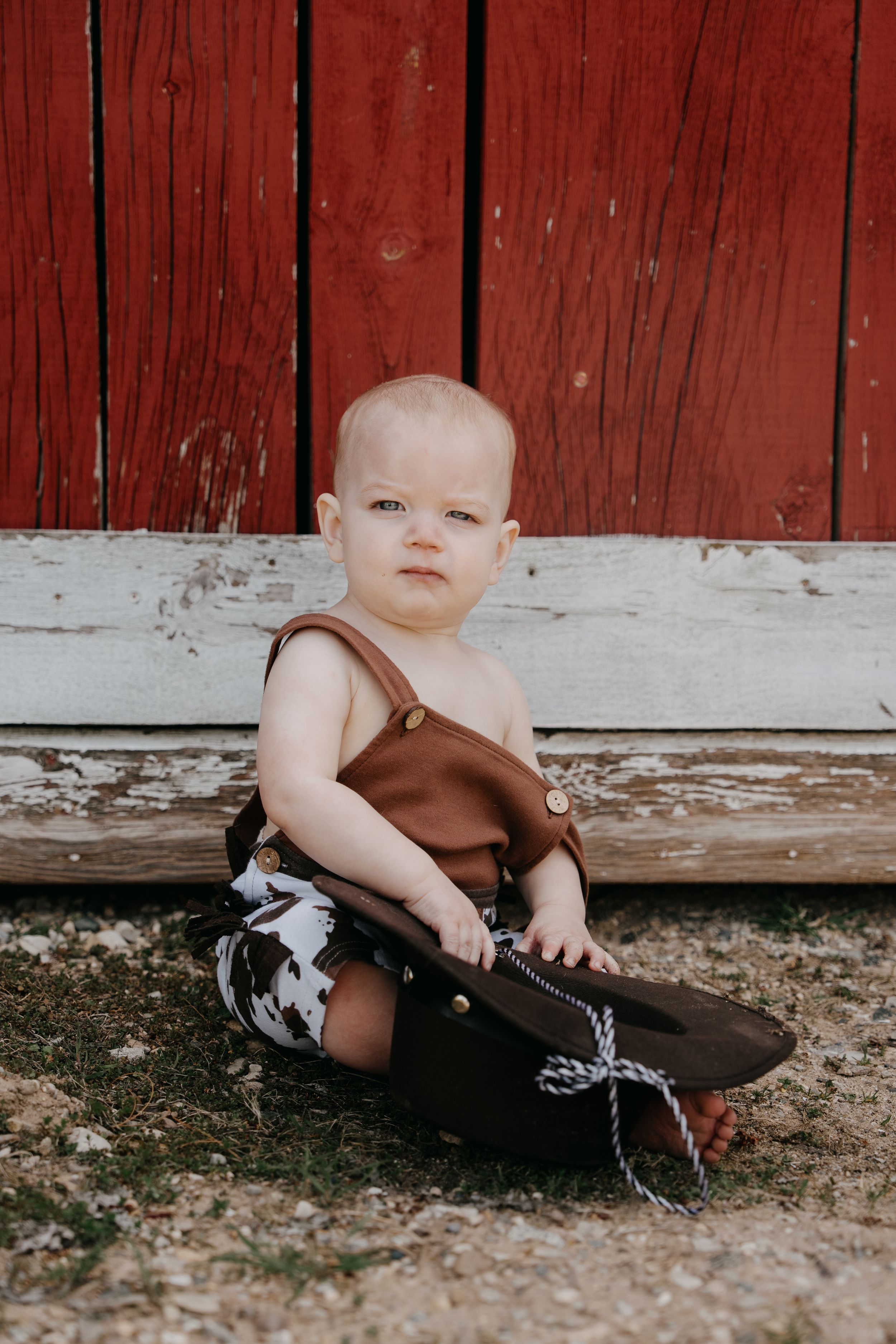 Baby in brown overalls sits by weathered white trim against red barn siding.