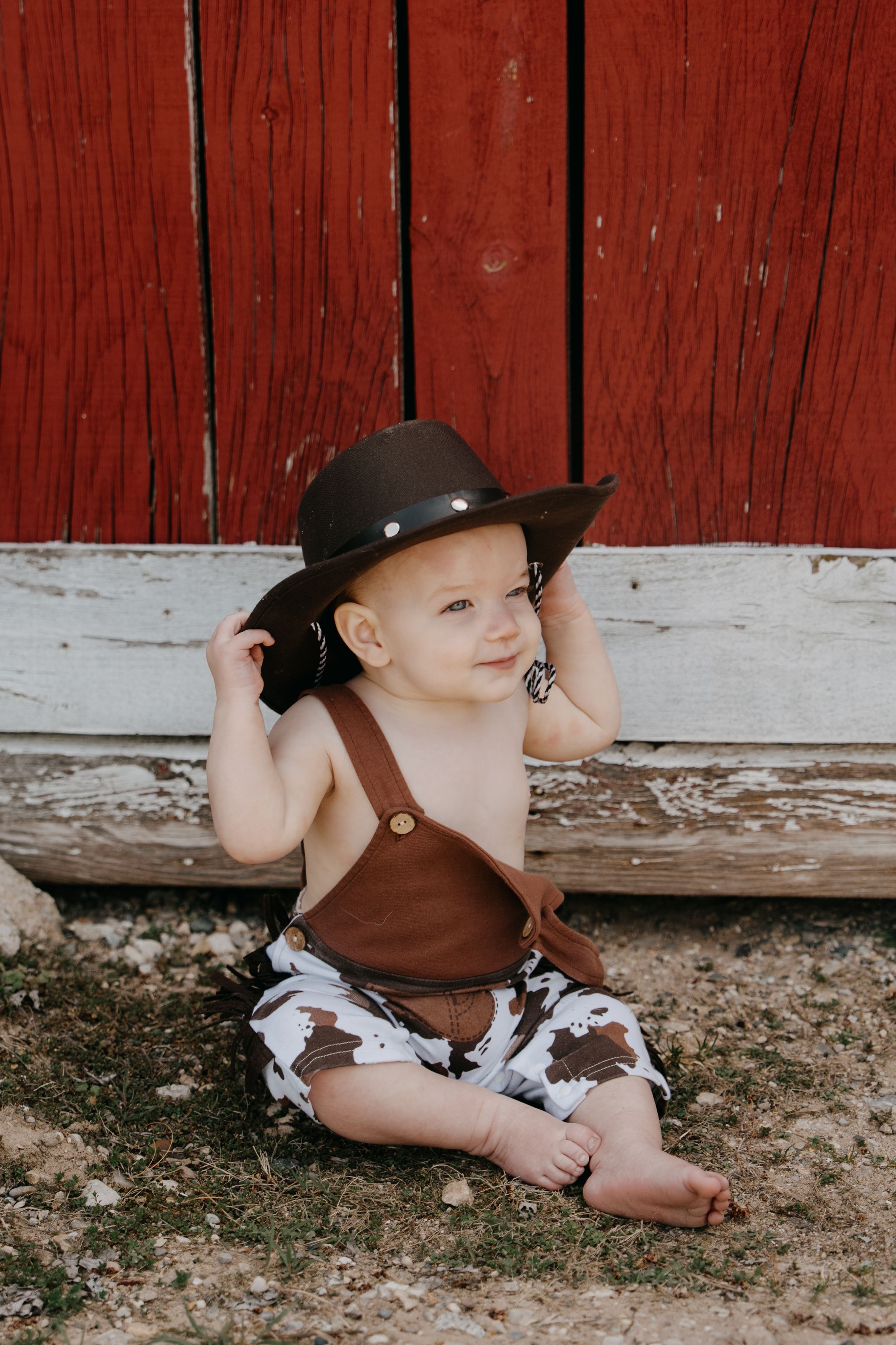 Small child in brown suspenders and cowboy hat sits against rustic red barn wall in countryside setting.