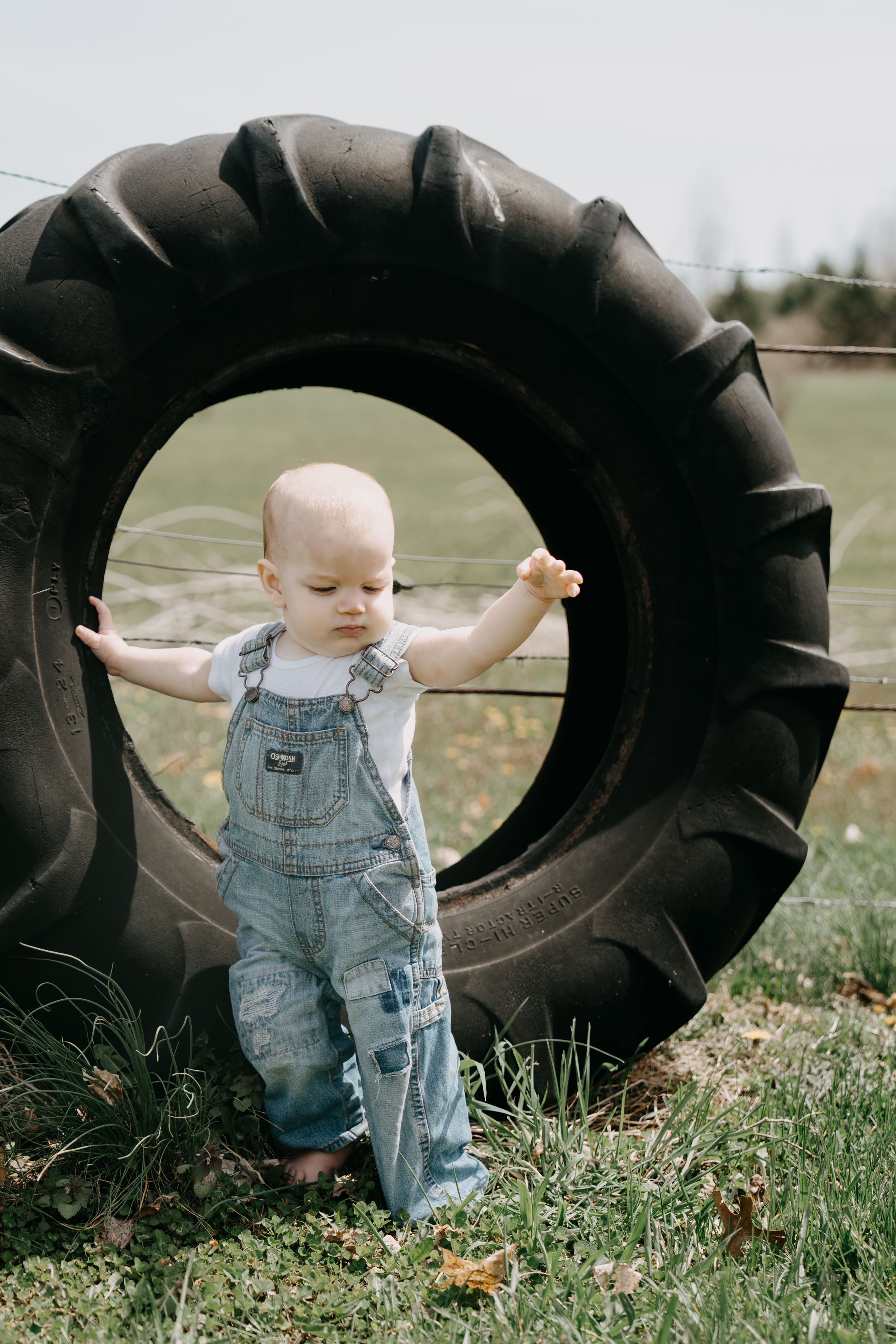 Small child in denim overalls stands beside large tractor tire in grassy field.