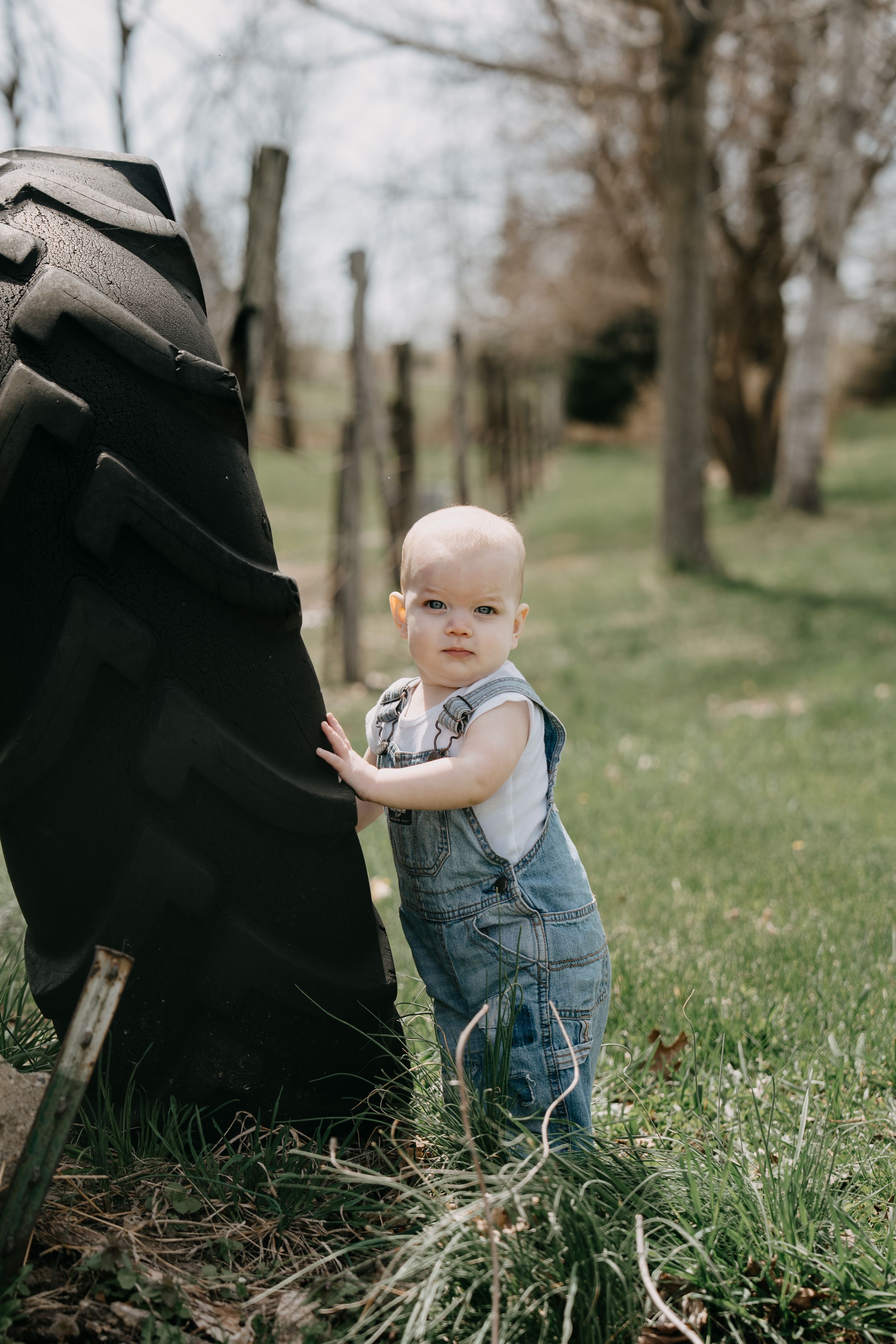 Small child in denim overalls stands outdoors near a black raincoat on a spring day.