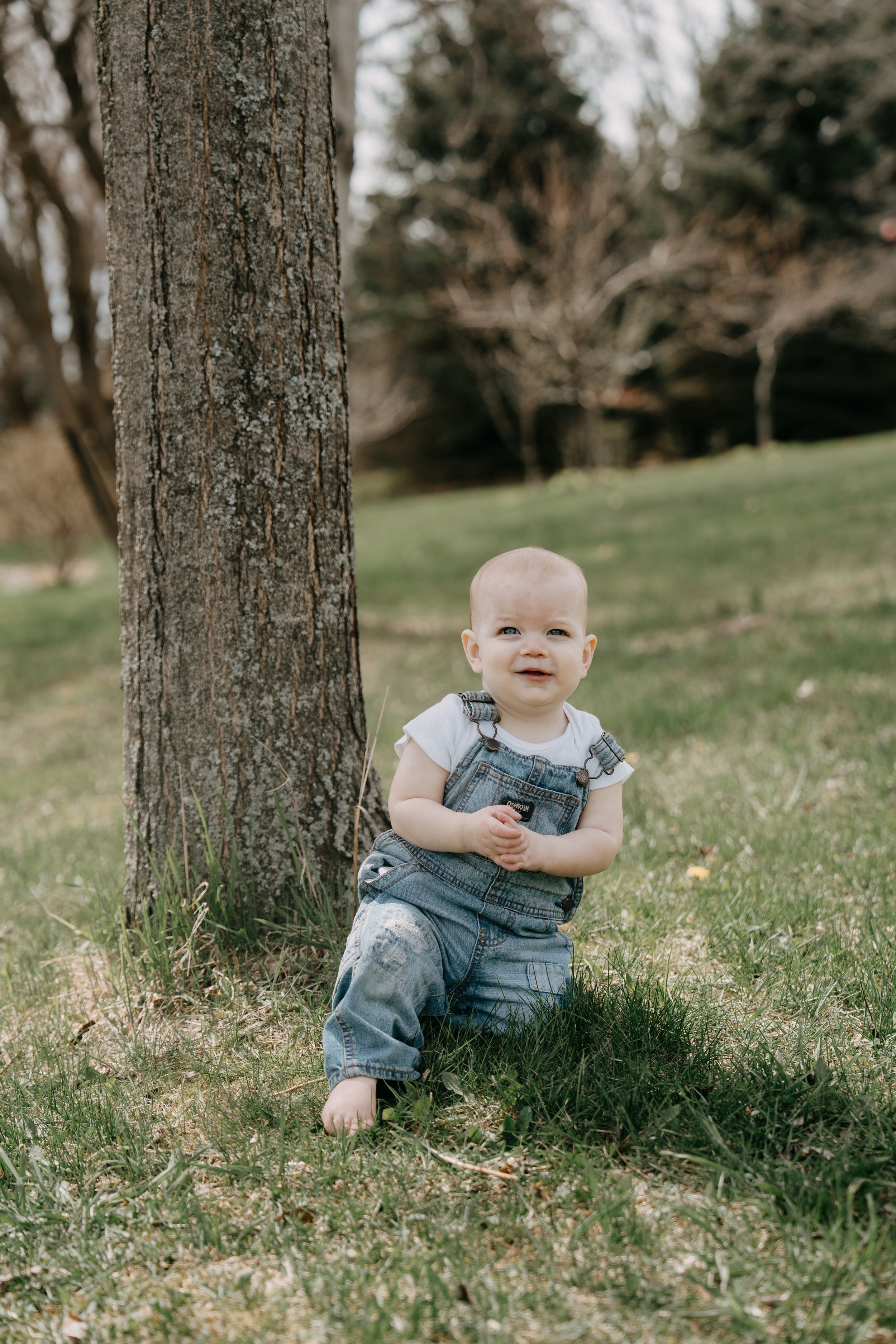 A smiling toddler in denim overalls sits against a tree trunk in a grassy outdoor setting.