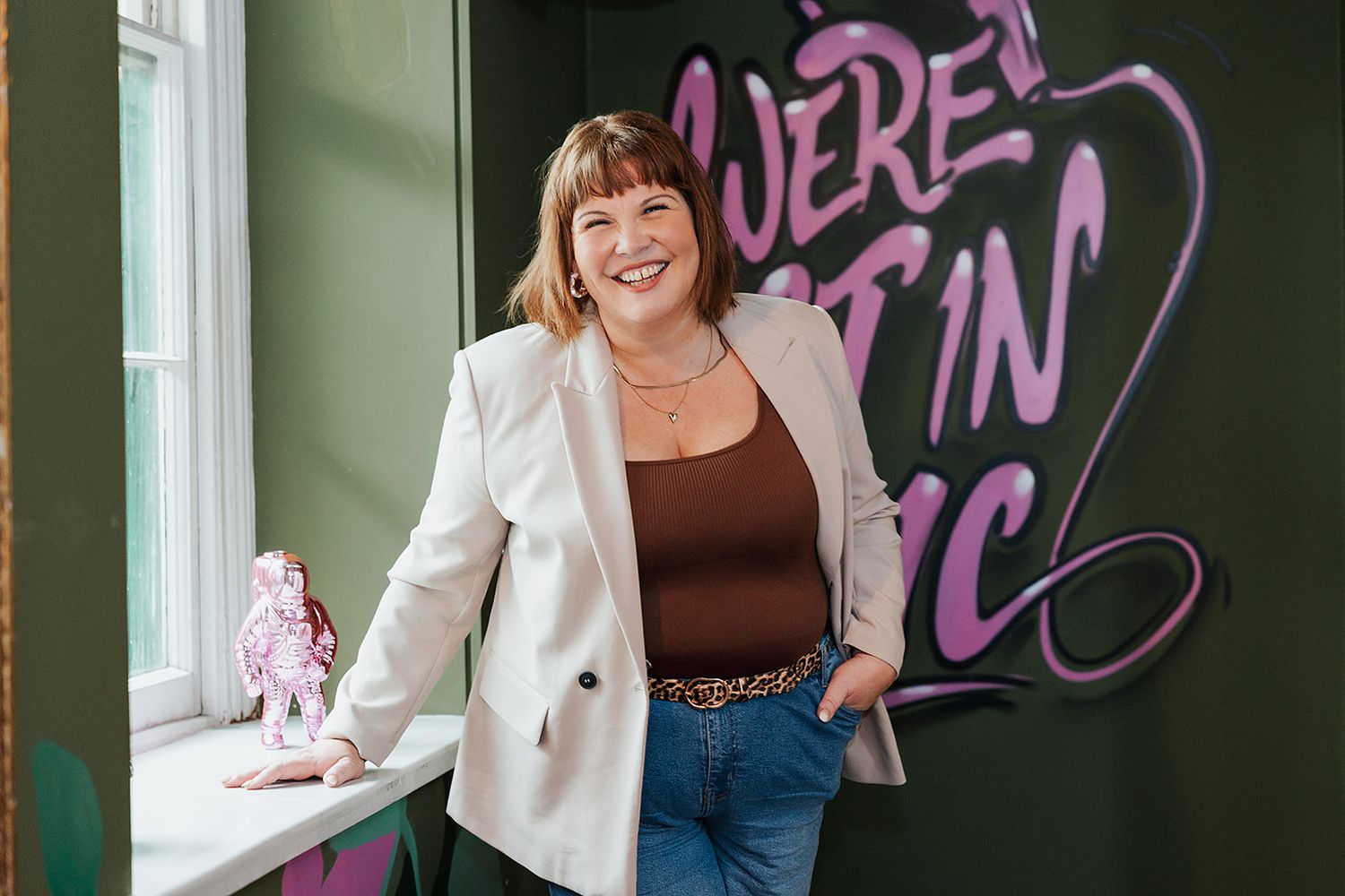 A person in a cream blazer and jeans stands smiling against a wall with neon text in a bright office space.
