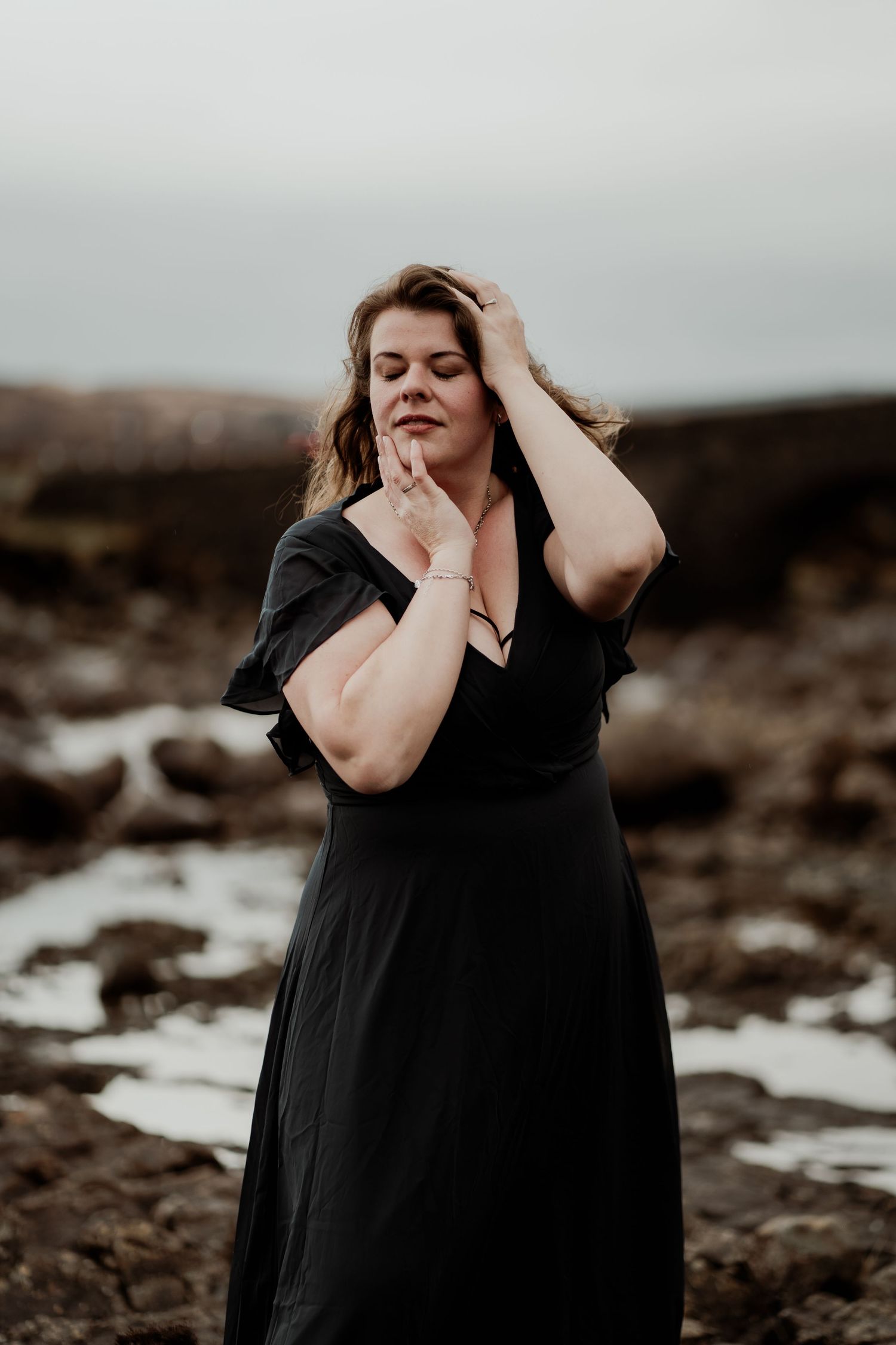 A woman in a black dress stands amid a moody landscape with patches of snow on the ground, running fingers through her hair.