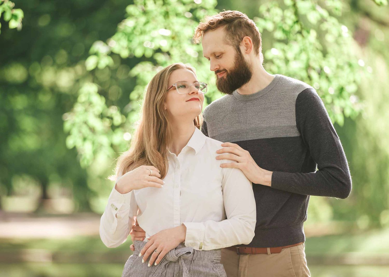Bride-to-be smiling while embraced by fiancé beneath greenery in Boston Public Garden Lagoon engagement session