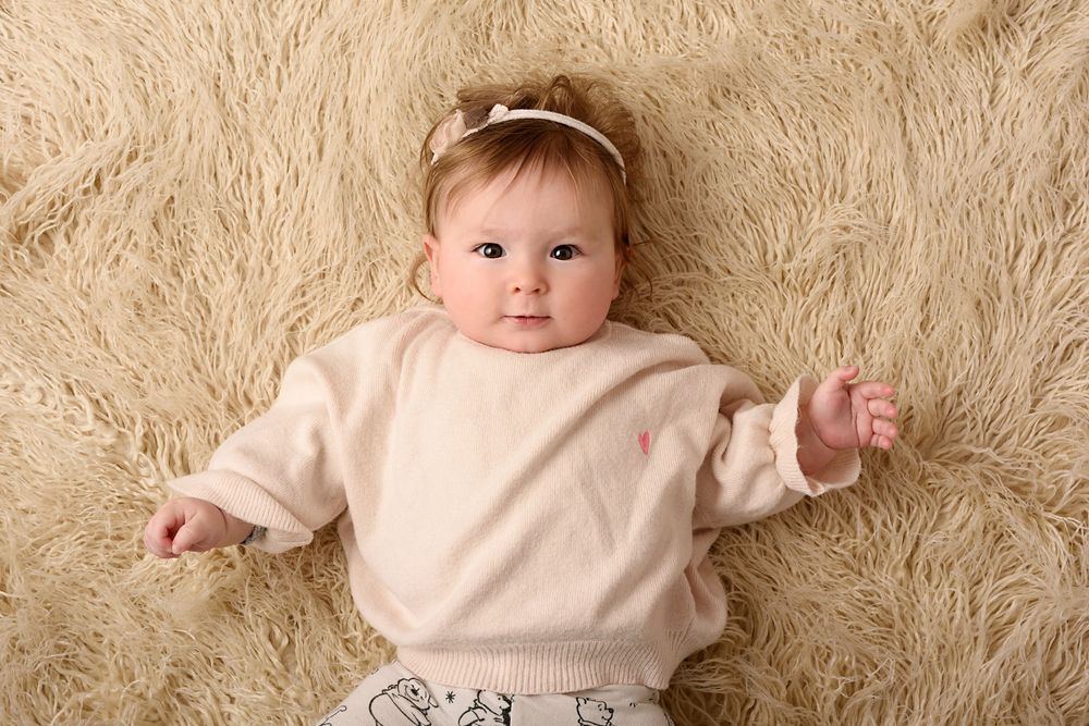 Baby wearing cream colored top poses on soft furry rug during portrait session.