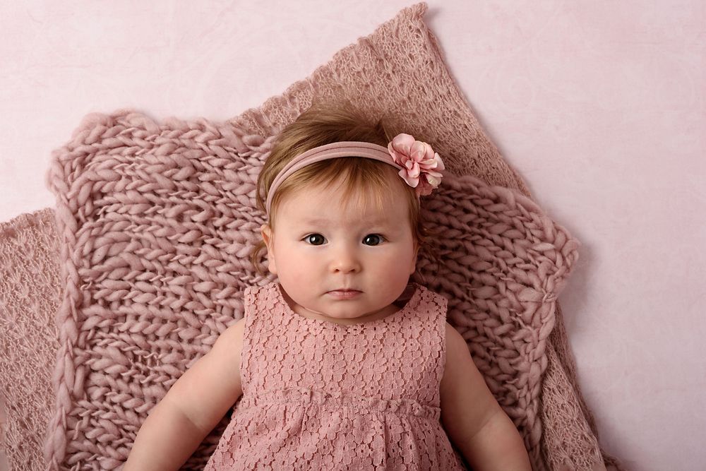 Baby in pink dress with flower hairband resting on textured blanket.