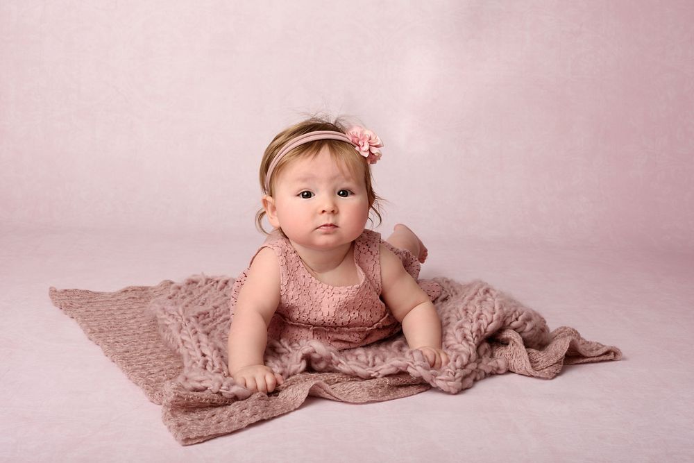 Baby in dusty rose dress poses on knitted blanket during  belly photo session.