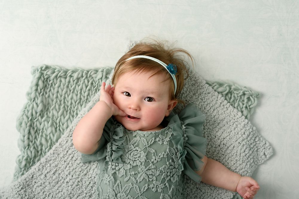 Baby in textured sage romper with headband resting on knitted blanket.