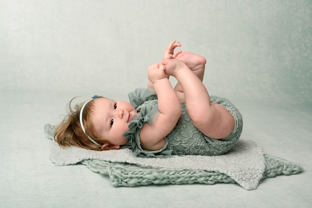 Baby in mint green outfit playfully grabbing feet during photo shoot.