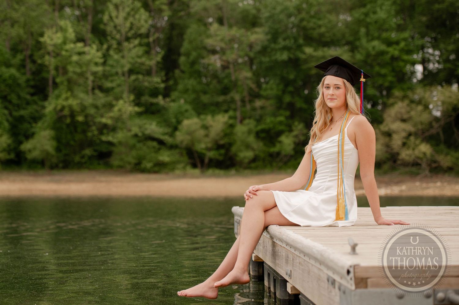 A graduate in a white dress and cap sits on a wooden dock by a serene lake surrounded by green trees.