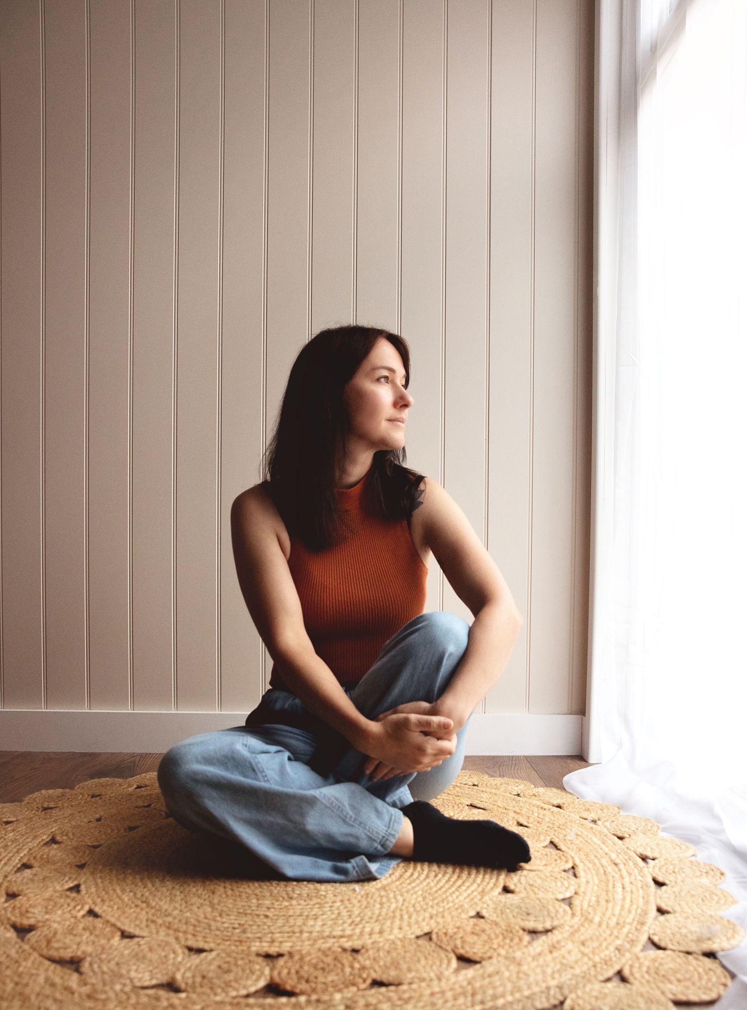 Person in rust-colored sleeveless top and jeans sits on round jute rug by window with natural lighting.