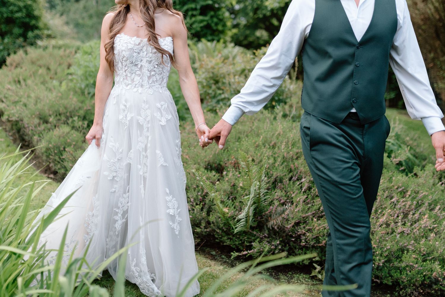 A couple in wedding attire holds hands while walking through lush green foliage at an outdoor venue.