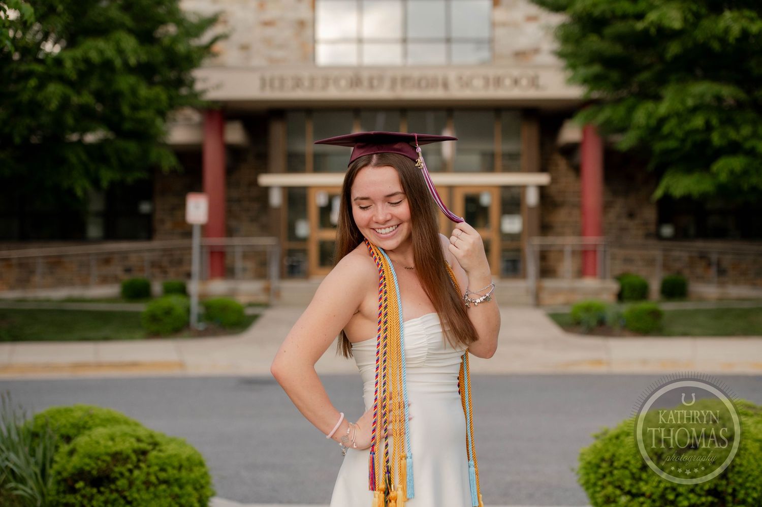 Graduate in white dress with honor cords poses smiling outside academic building entrance on graduation day.