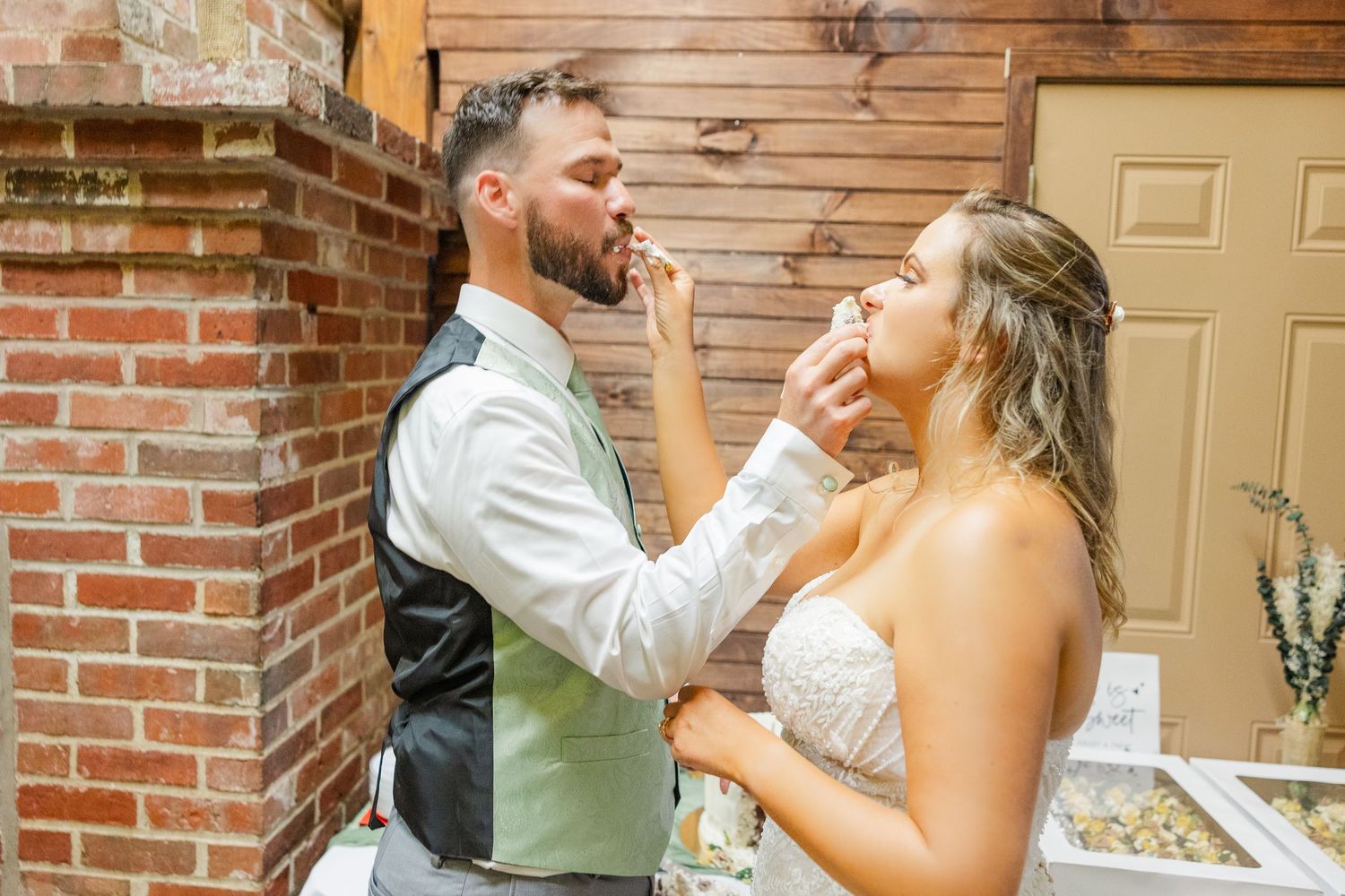 Joyous newlyweds feed each other pieces of wedding cake in front of exposed brick wall at reception.