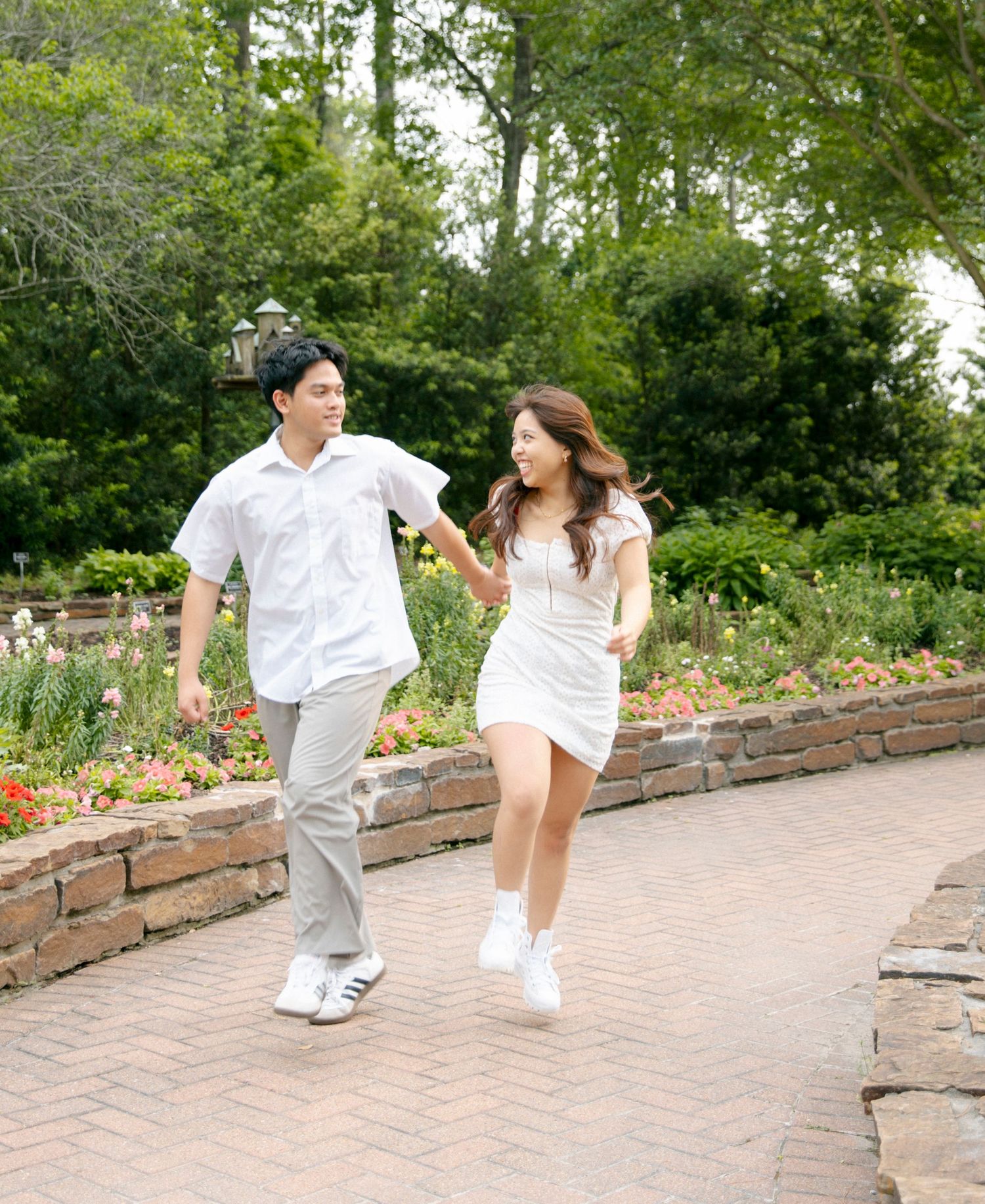 Happy couple running joyfully together along a brick pathway in a lush garden setting with flowers and greenery.
