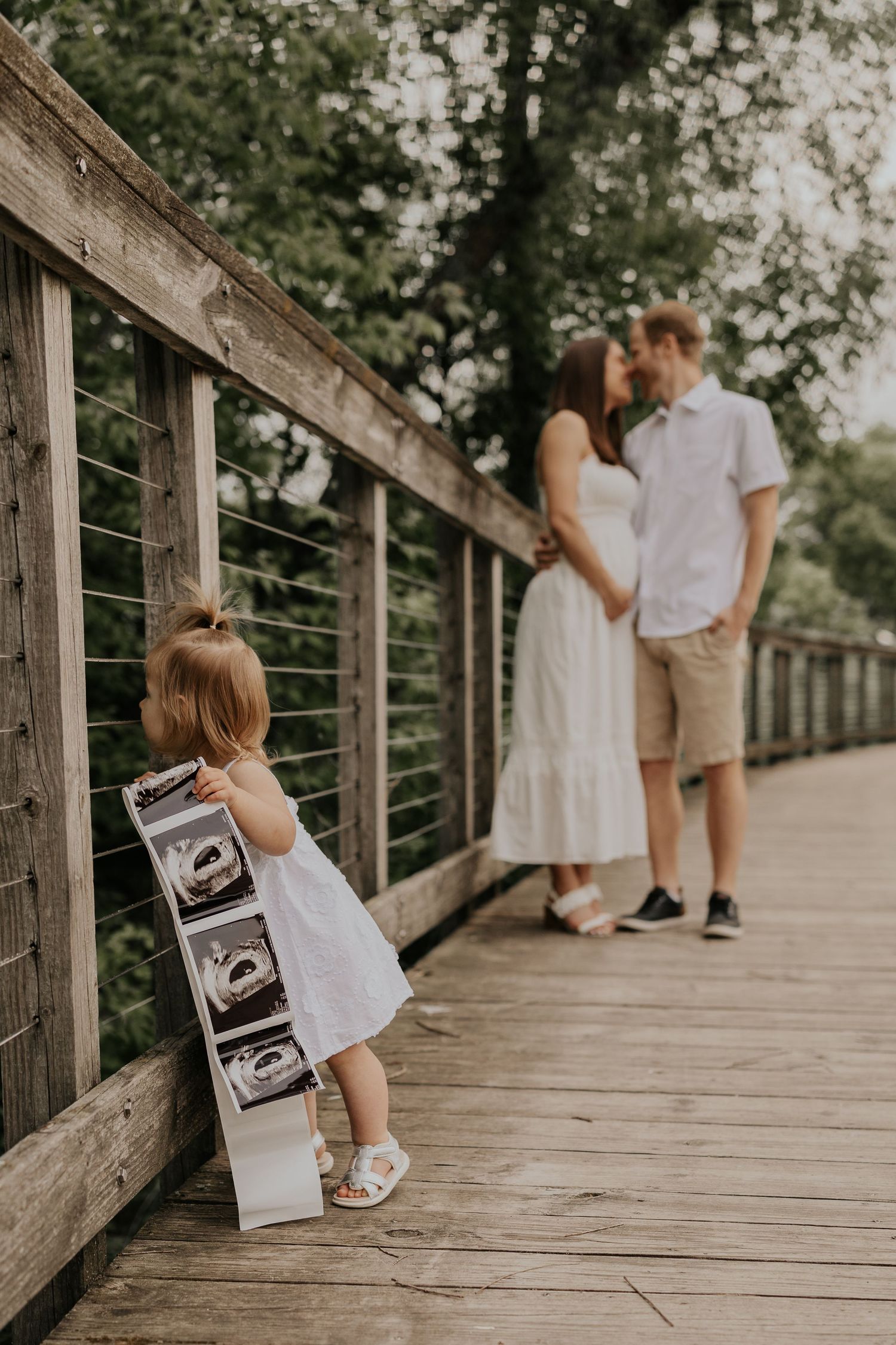 A toddler in a white dress holding ultrasound photos on a wooden bridge while a couple stands together in the background.