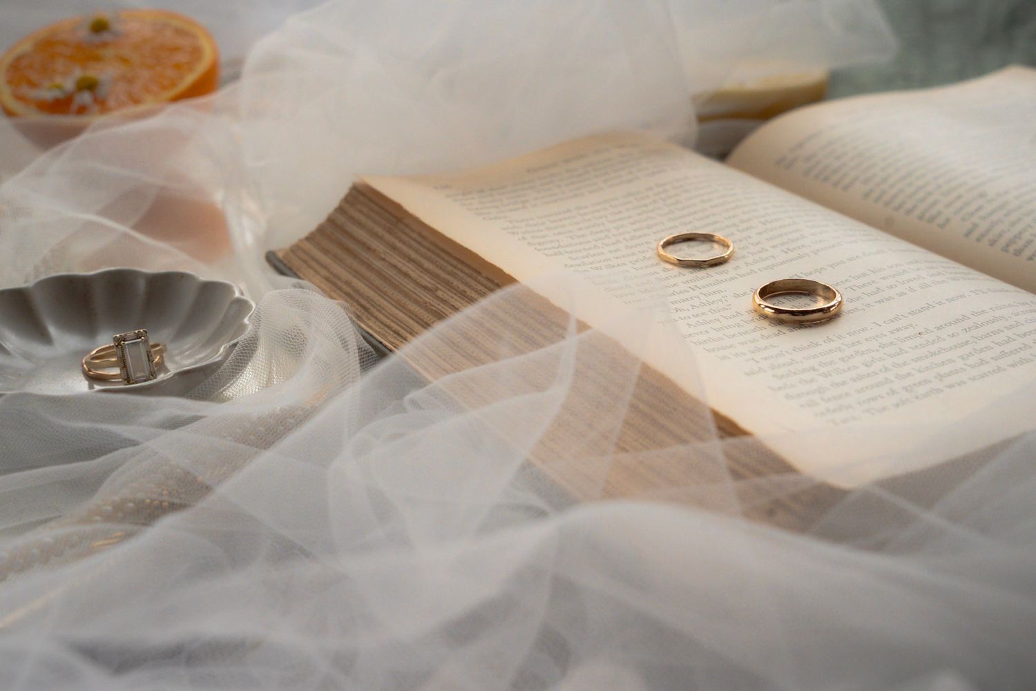 Two wedding rings rest on an open book page with soft white tulle fabric creating a romantic composition. Intimate elopement photographer