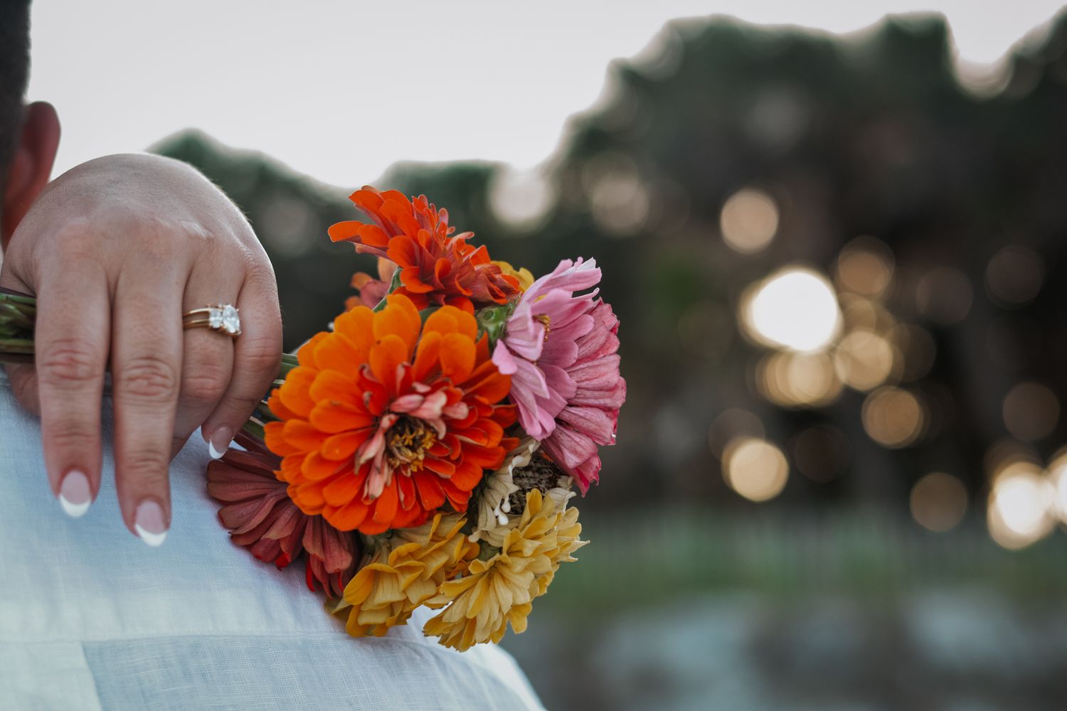 Hand displaying wedding ring with colorful zinnia bouquet in sunset light.