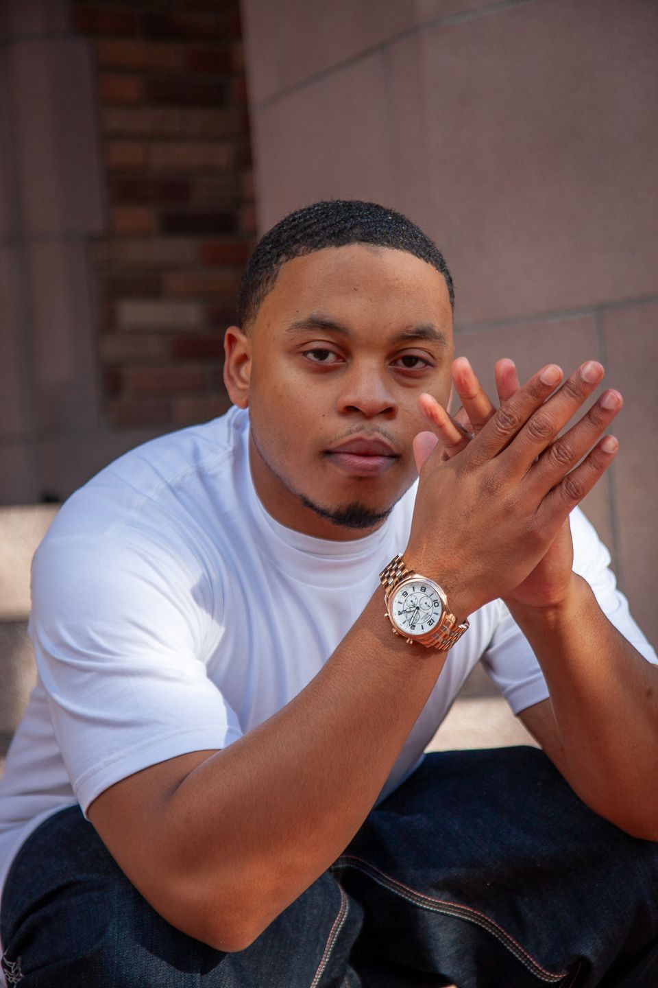 A person in a white t-shirt with a gold watch poses with clasped hands while sitting outdoors against a brick wall.