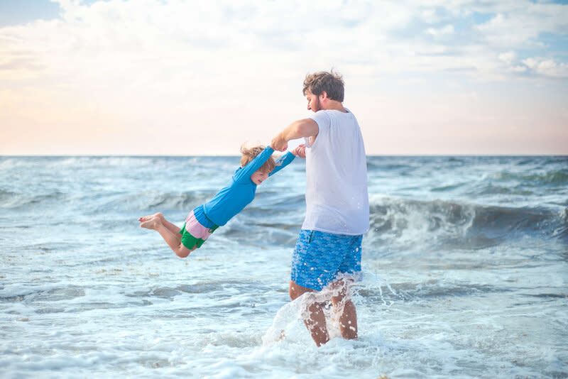Adult in white shirt and blue shorts spins child playfully at beach shore with ocean waves in background at sunset.