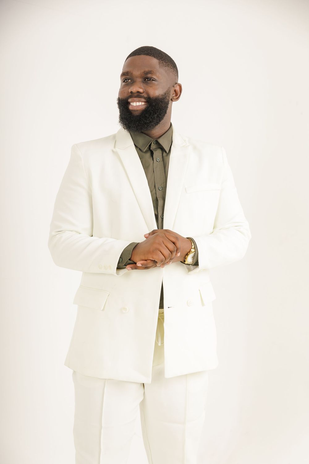Smiling person wearing a crisp white suit with olive dress shirt poses against light background with clasped hands.
