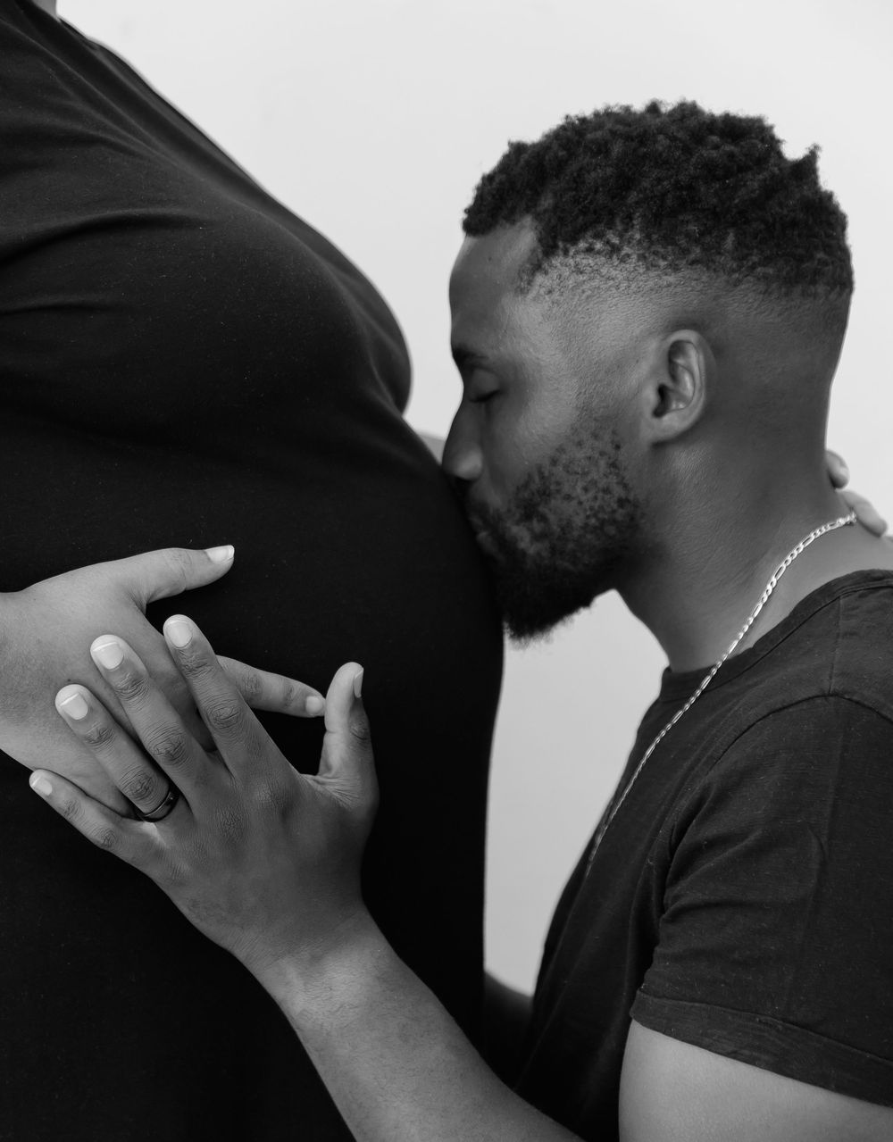 A tender black and white portrait of a person kissing a pregnant woman's belly while gently holding her stomach.