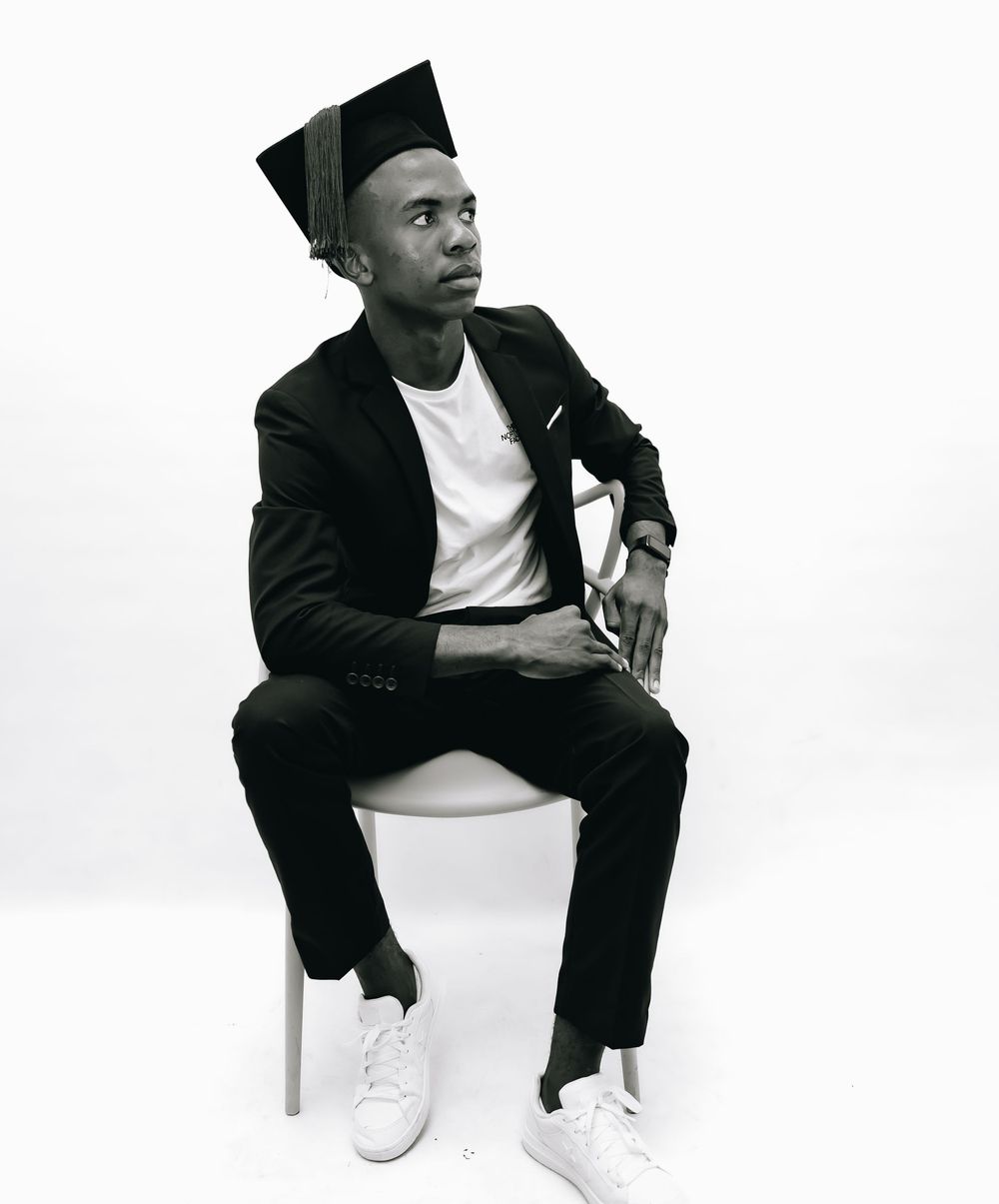 A person in a black suit and white sneakers sits on a chair against a white background in a minimalist portrait.